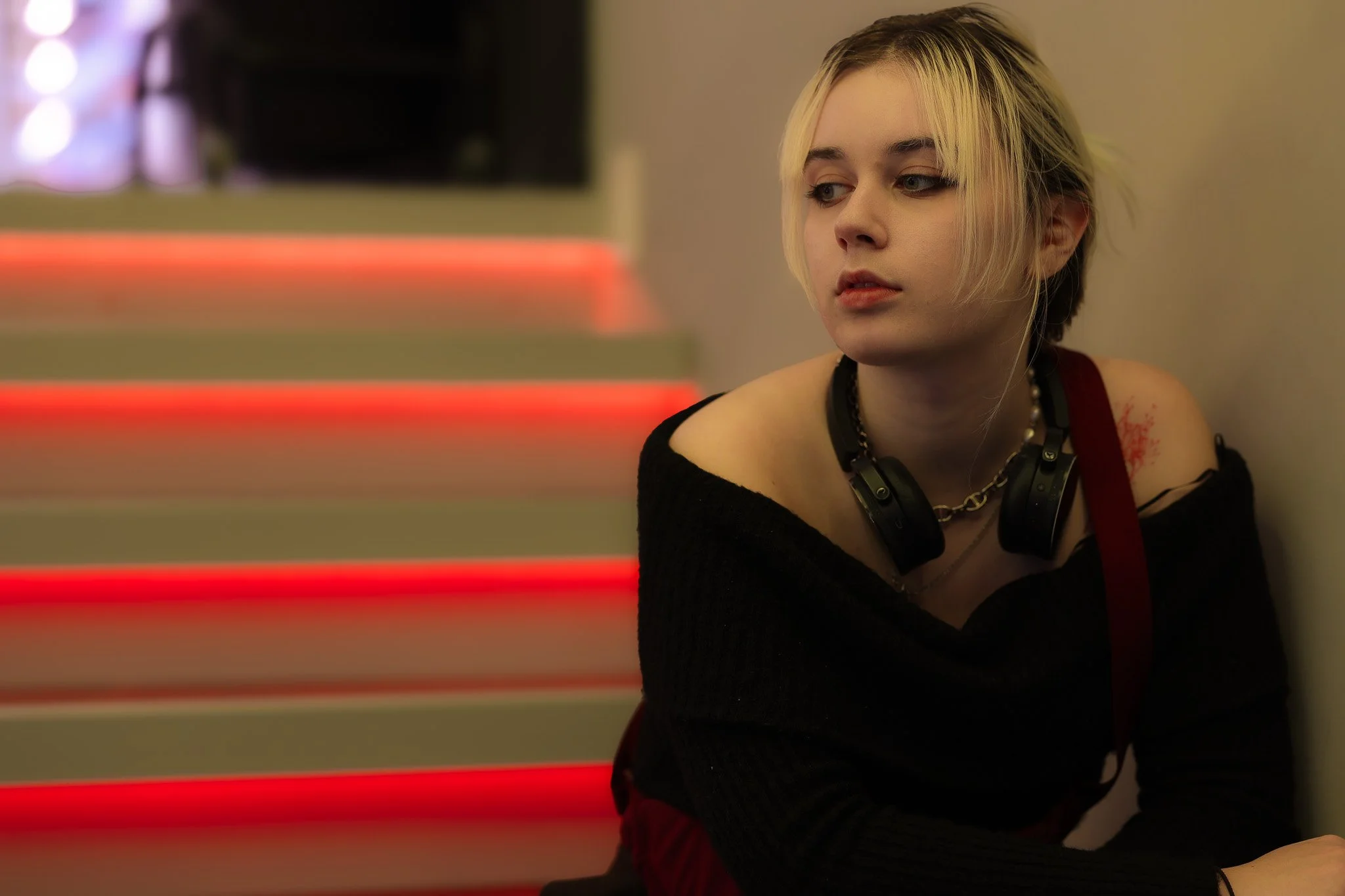 A young woman with short blonde hair, wearing a black off-shoulder top and a silver necklace, sits indoors with a neutral expression. She has headphones around her neck and is looking to her right. The background features horizontal red and white str