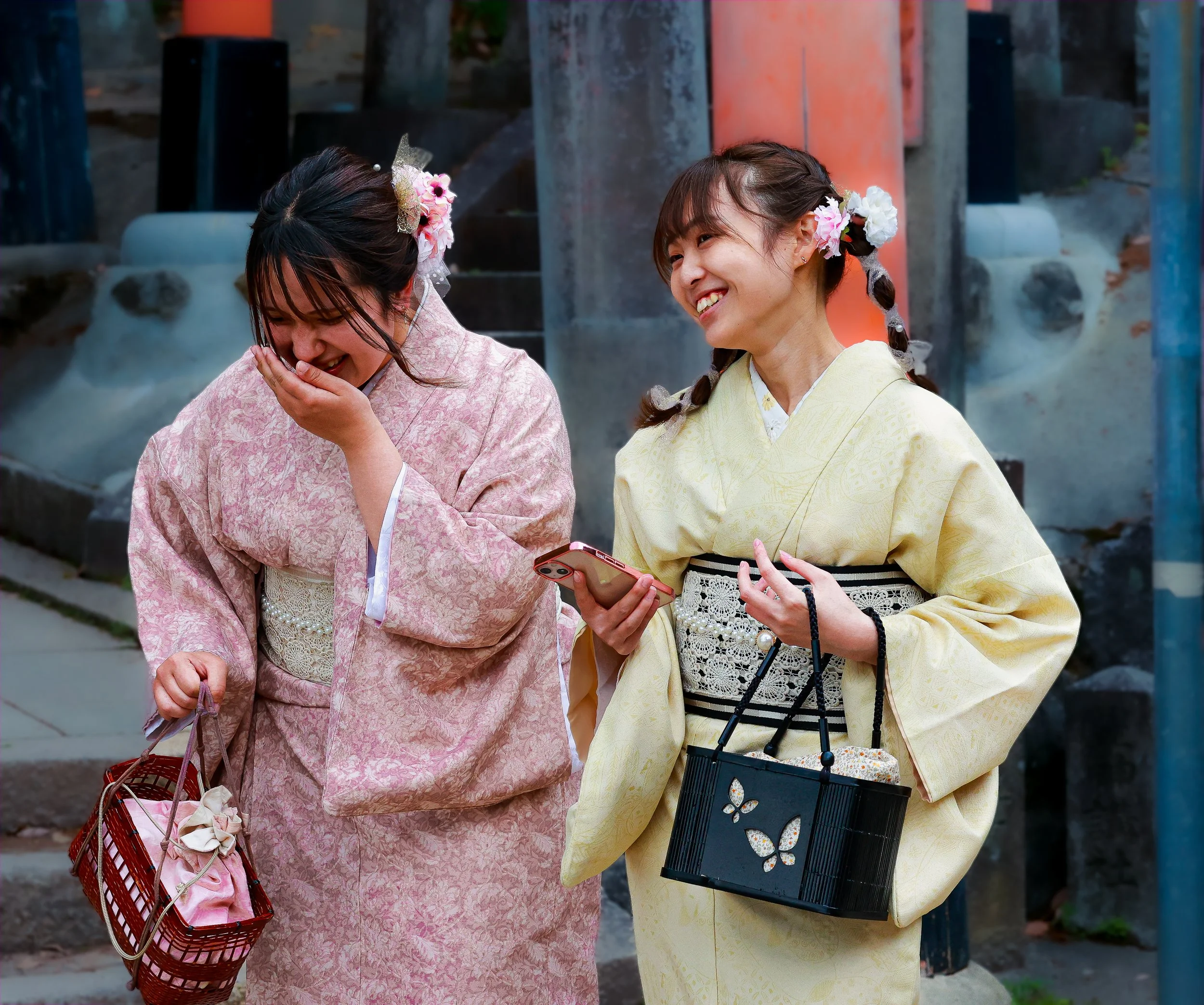 Two young women dressed in traditional Japanese kimonos, smiling and laughing together. One woman is holding a small red basket, and the other is holding a smartphone. They have flowers in their hair and are standing outdoors near a stone or concrete structure.