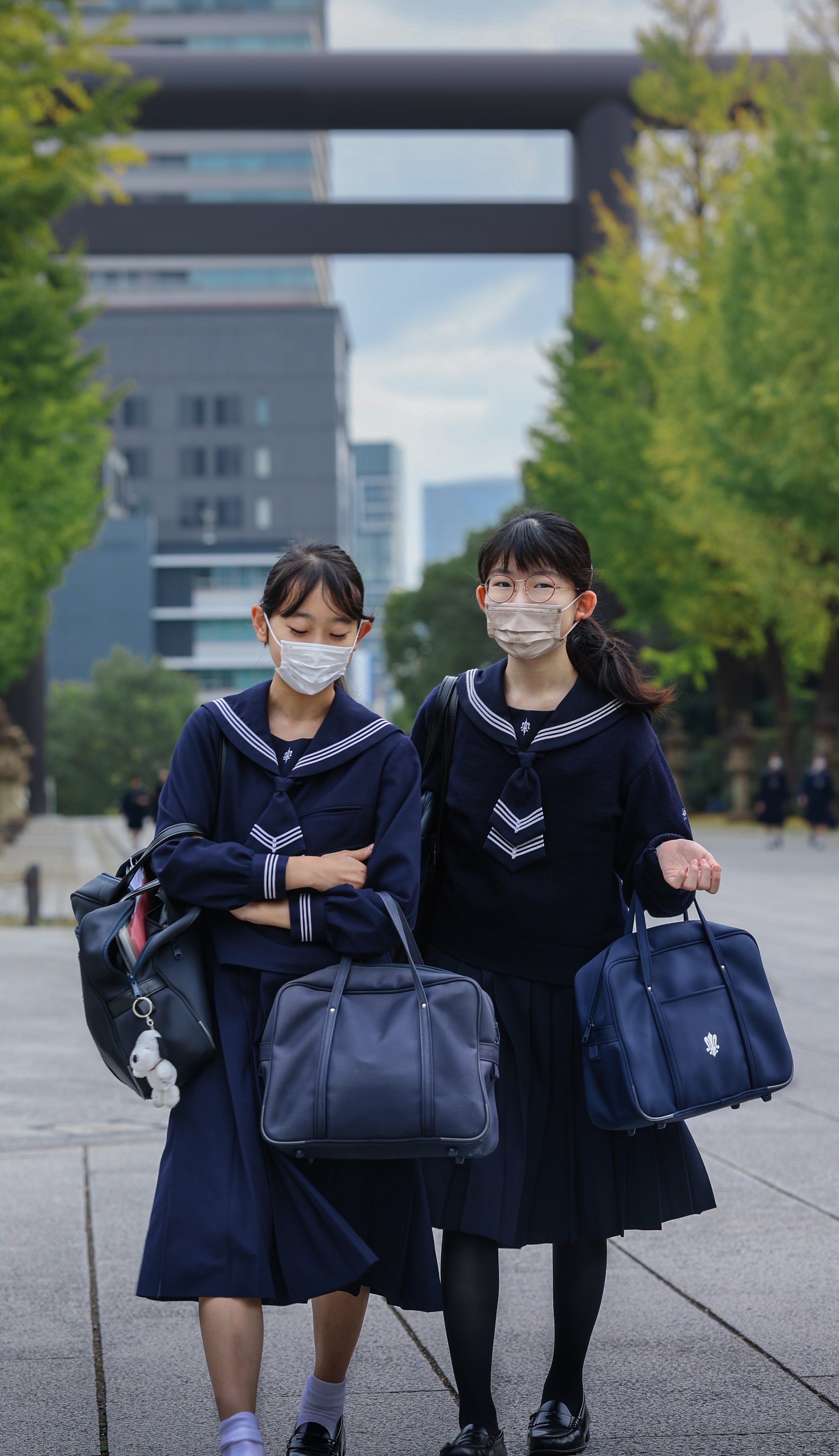Two schoolgirls wearing navy blue uniforms and face masks are walking together with school bags in hand on a city street lined with trees and modern buildings.
