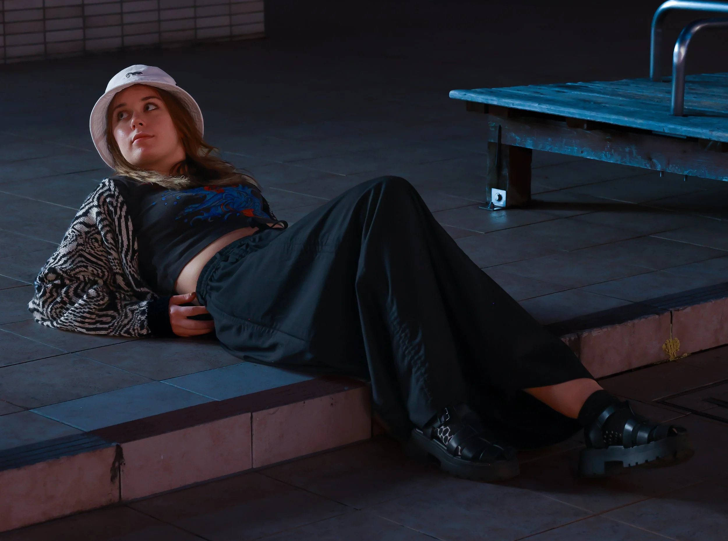 A young woman lying on a tiled step at night, wearing a white bucket hat, a patterned black and white cardigan, dark pants, and black shoes.