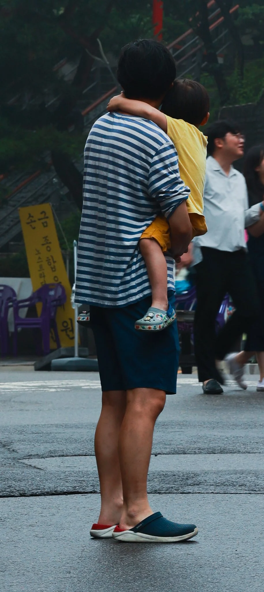 A man wearing a striped blue and white shirt, black shorts, and crocs stands on a crosswalk holding a child in a yellow shirt and patterned sneakers. The background shows other people walking and a yellow sign with black writing.