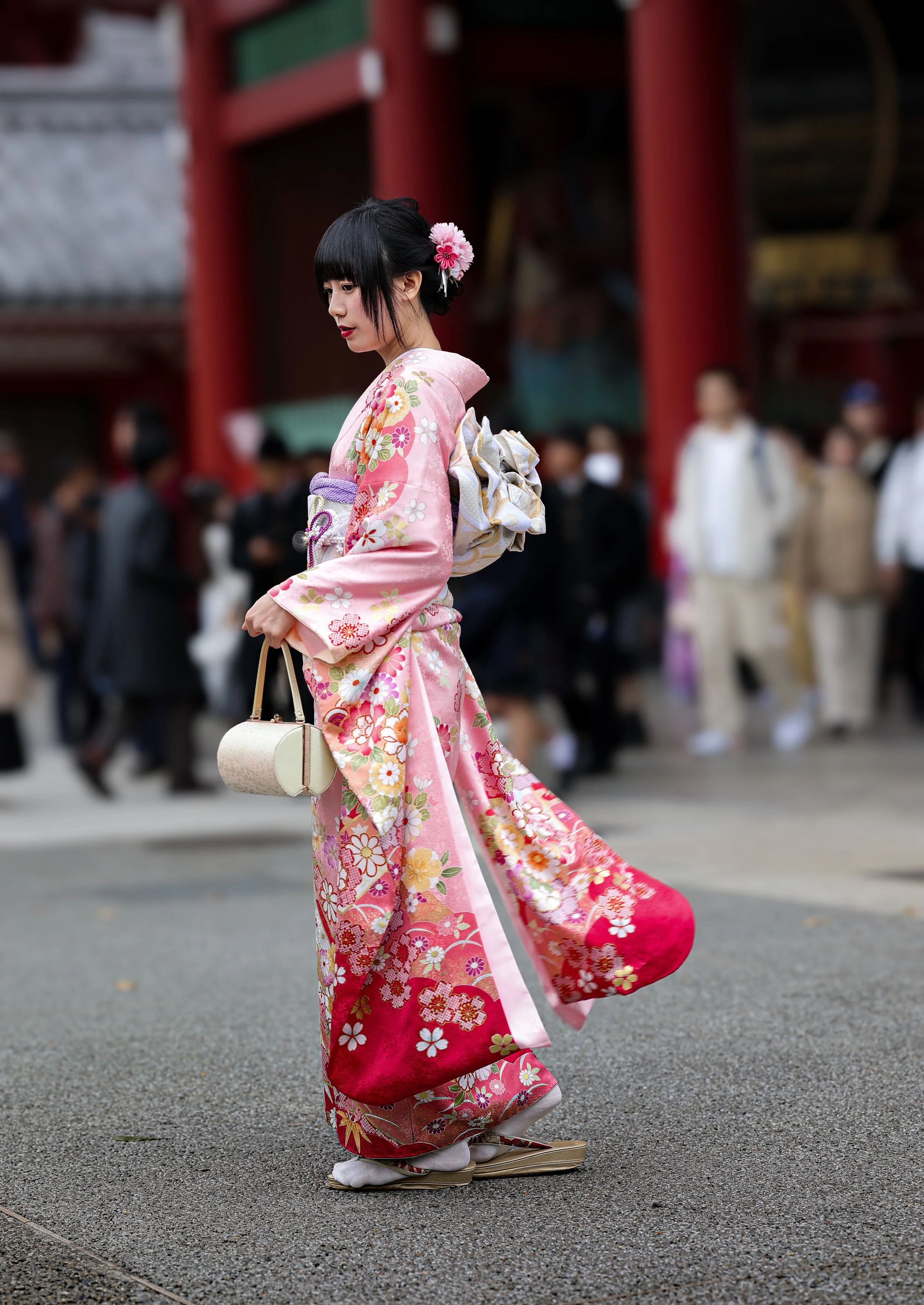A young woman in a pink kimono with floral patterns is walking on a street. She has short black hair adorned with pink flowers and is carrying a small white handbag. The background shows a crowd of people and traditional architecture.