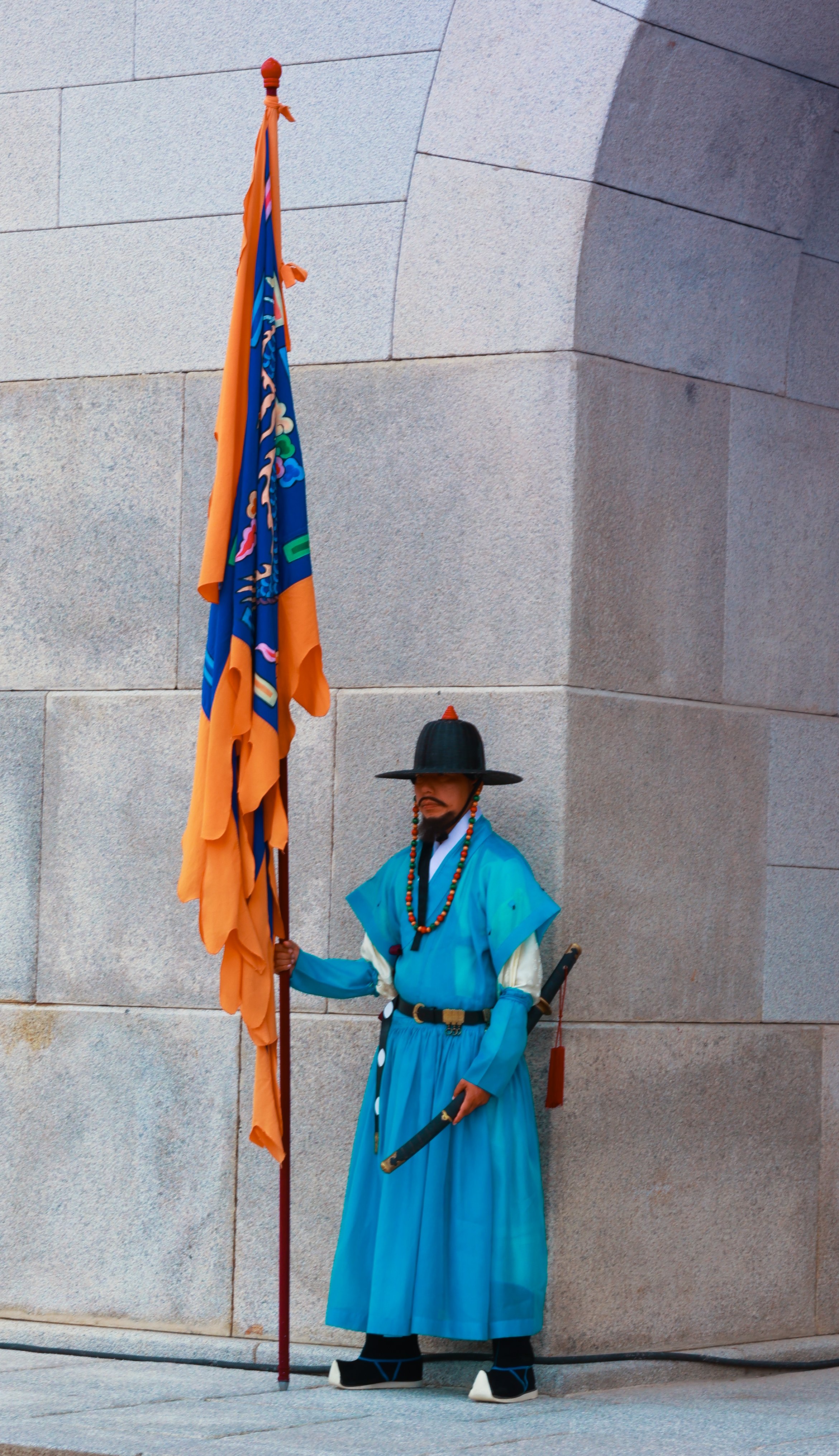 A man dressed in traditional Korean guard attire, standing next to a stone wall and holding a large multi-colored flag. He is wearing a blue hanbok, black hat, and traditional footwear.