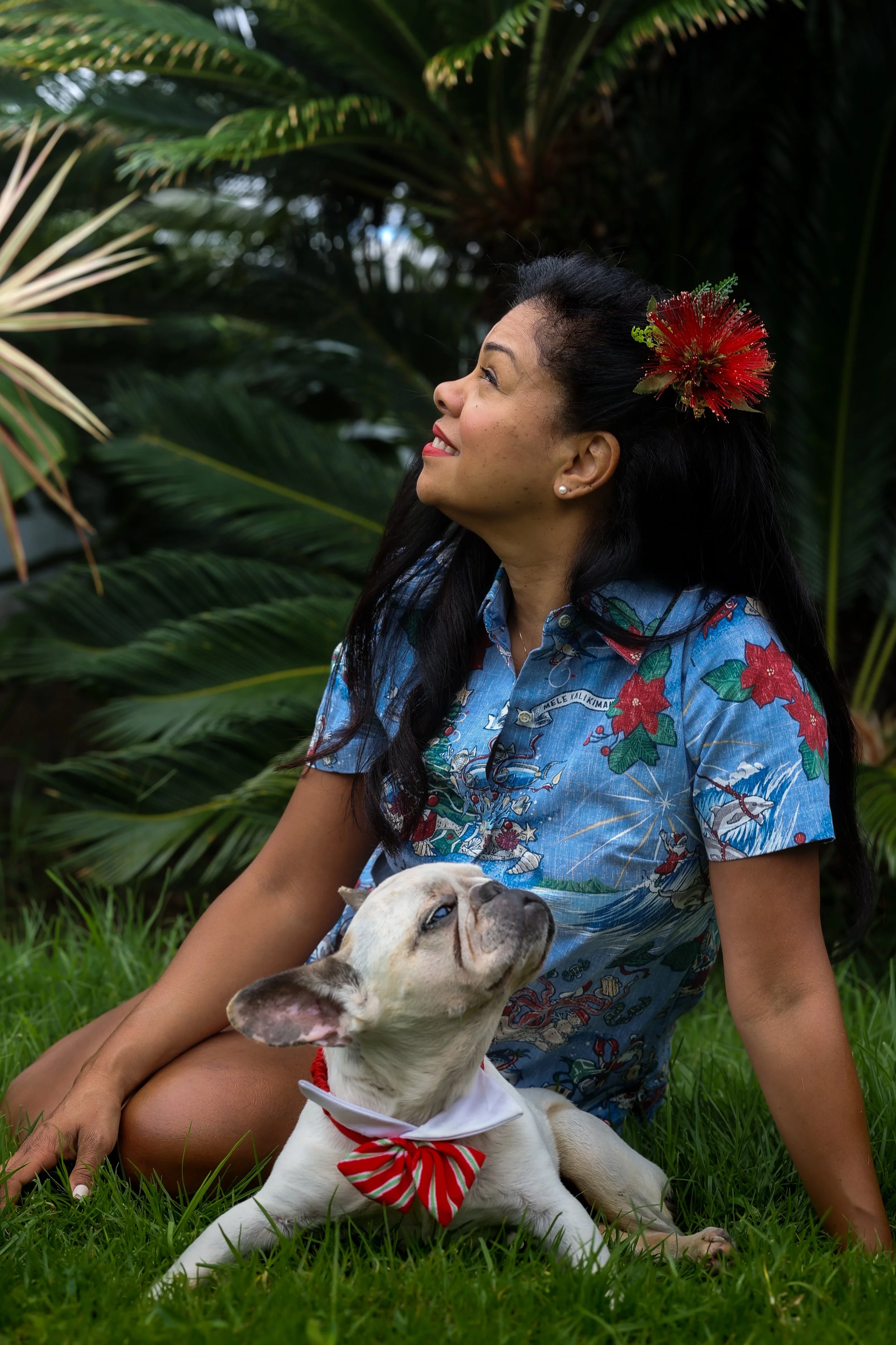 A woman with dark hair and a red flower in her hair, sitting on grass in front of green foliage, smiling and looking up, holding a white dog with black spots and a red and white striped bow tie.