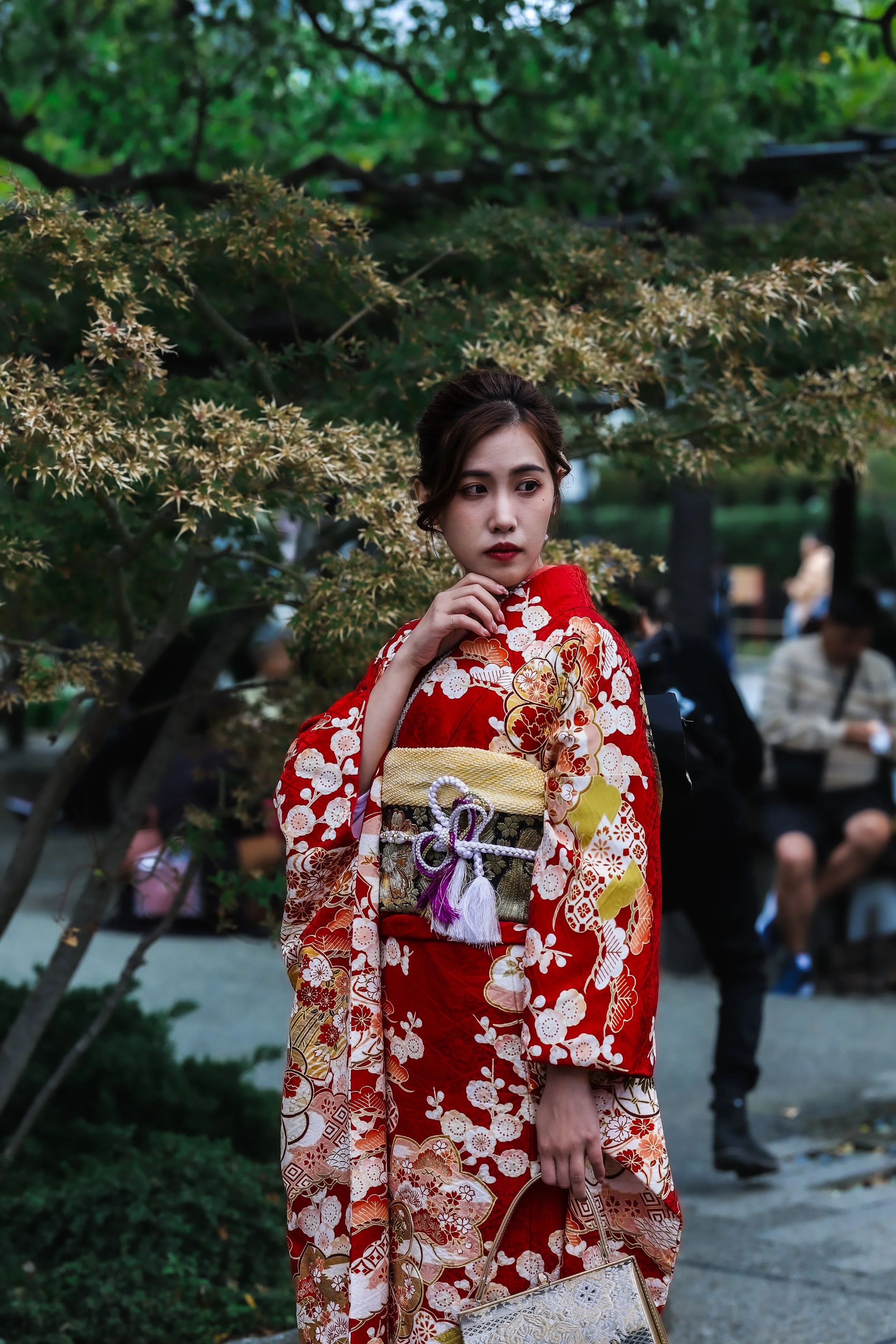 A young woman in a red kimono with white floral patterns, holding a patterned handbag, standing outdoors near autumn leaves and trees, with blurred people in the background.
