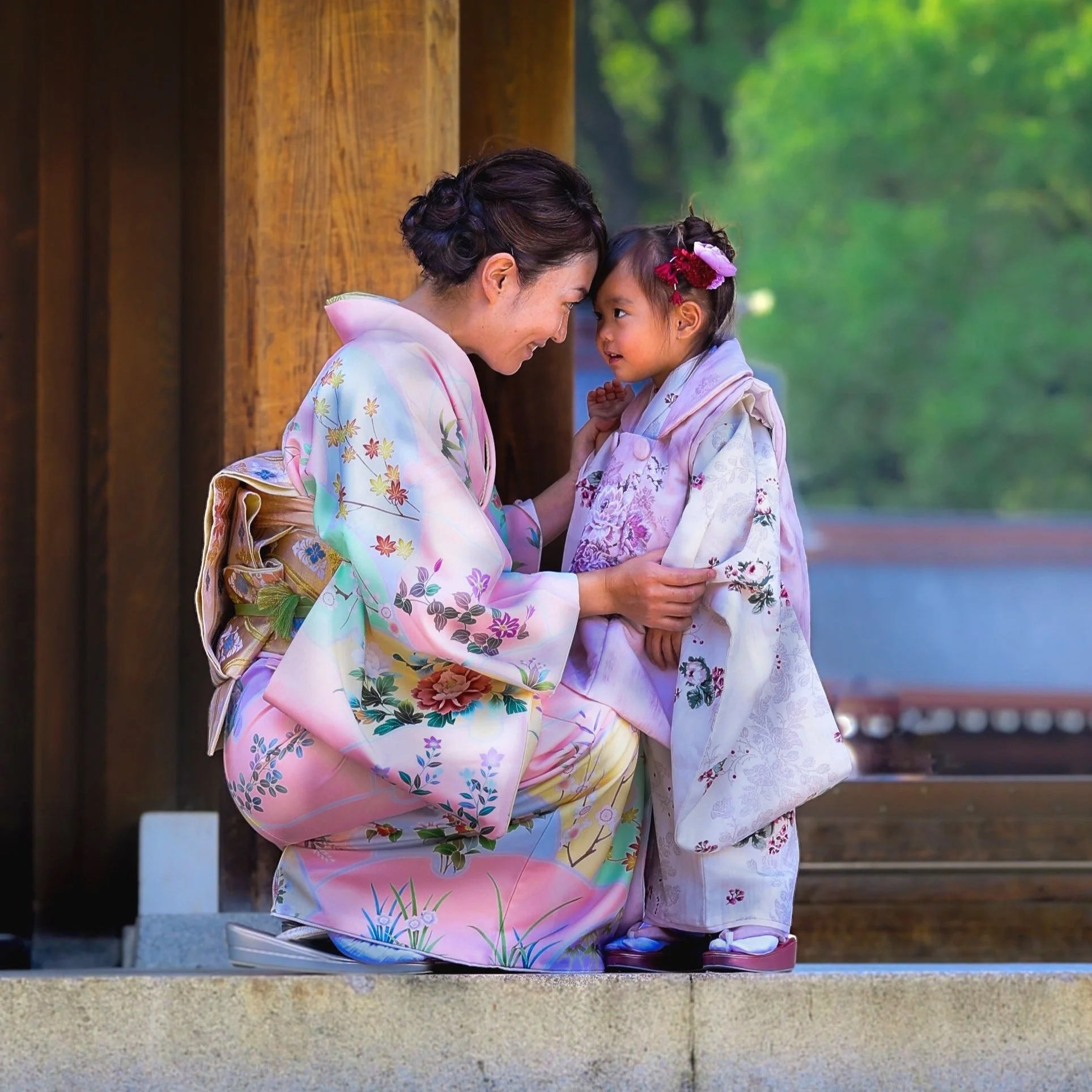 A woman and a girl in traditional Japanese kimonos touching foreheads and smiling outdoors, with a wooden structure and green trees in the background.