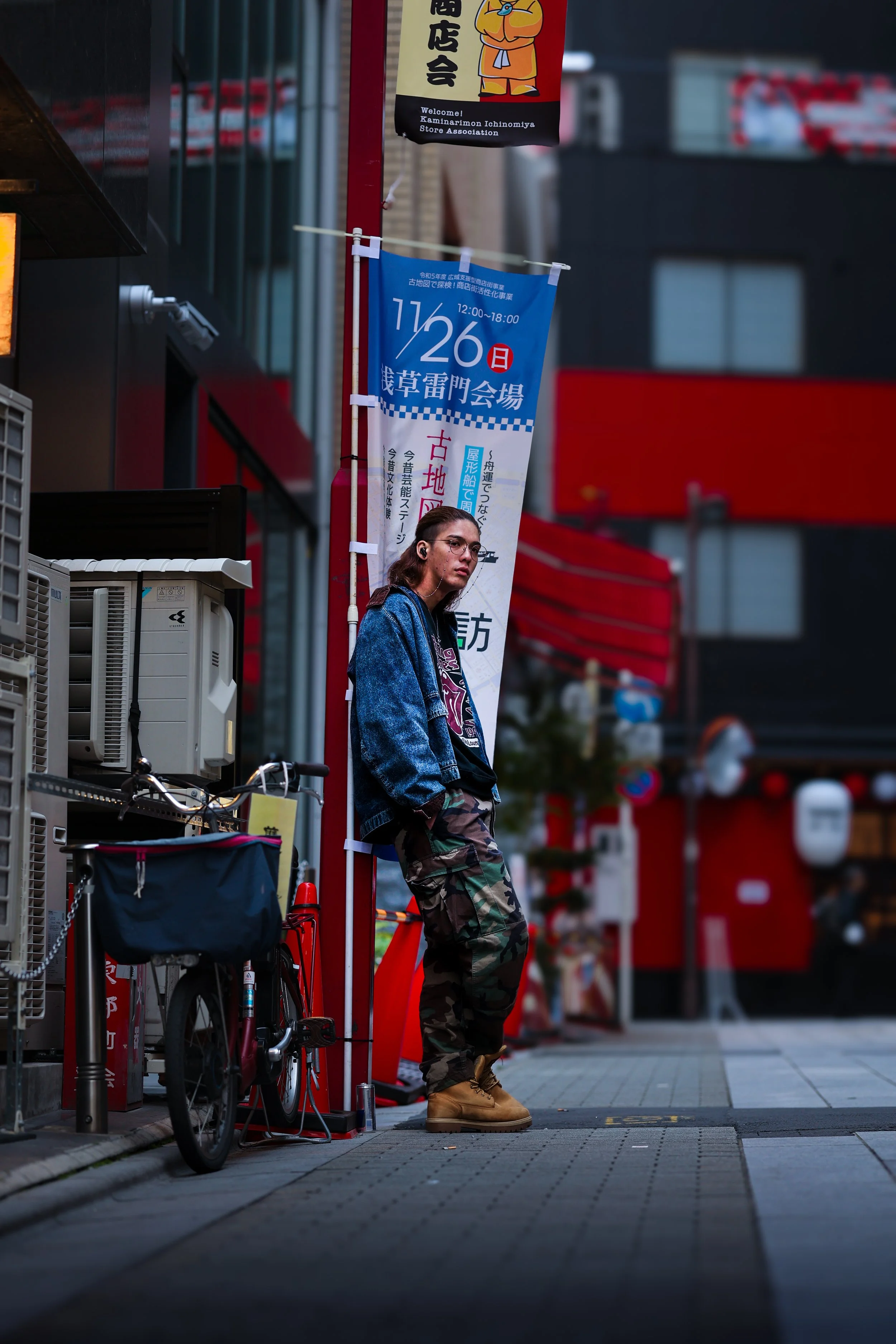 A young person with long hair, glasses, and wearing camouflage pants, a denim jacket, and tan boots, standing on a city sidewalk next to colorful vertical banners with Japanese characters, bicycles, and air conditioning units visible nearby.