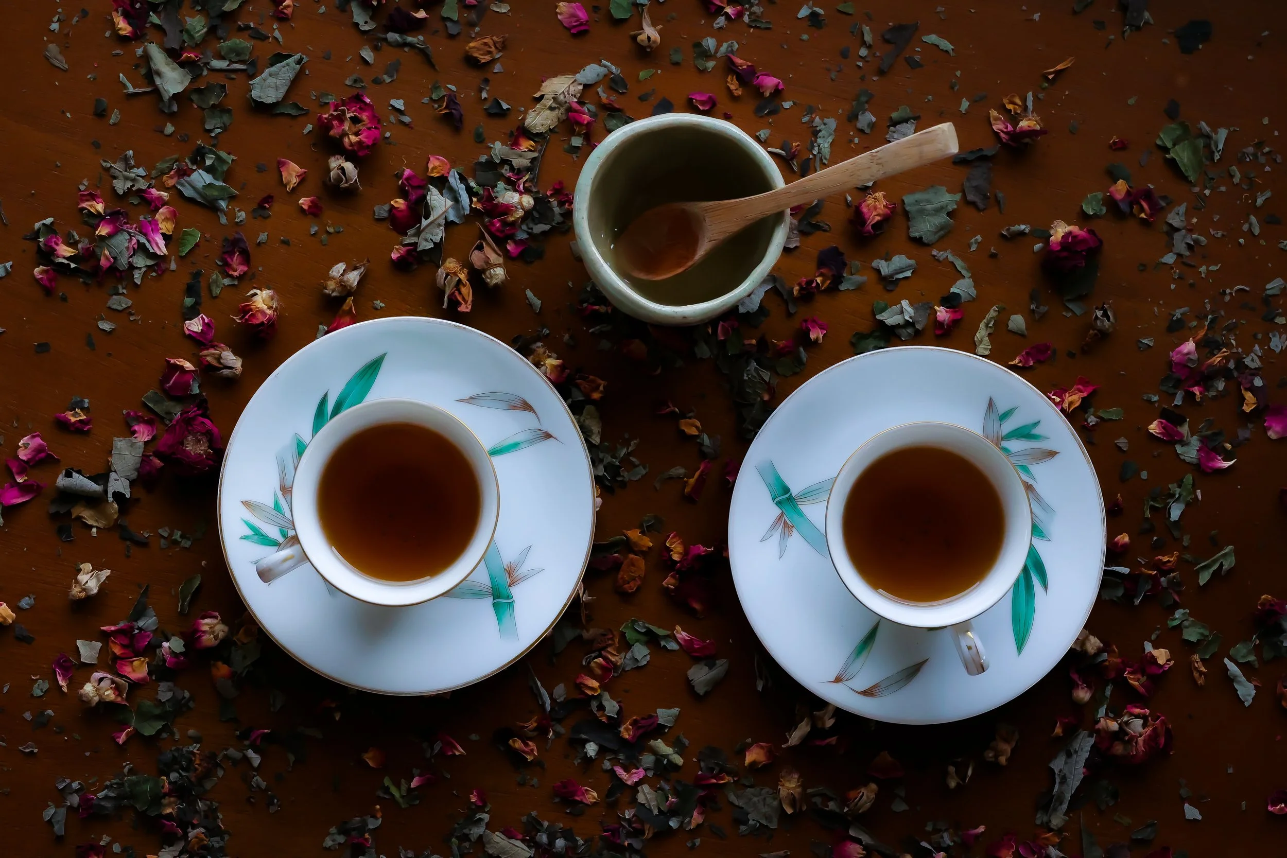 Three white porcelain cups filled with dark tea on saucers, surrounded by scattered dried flower petals and leaves on a wooden surface.
