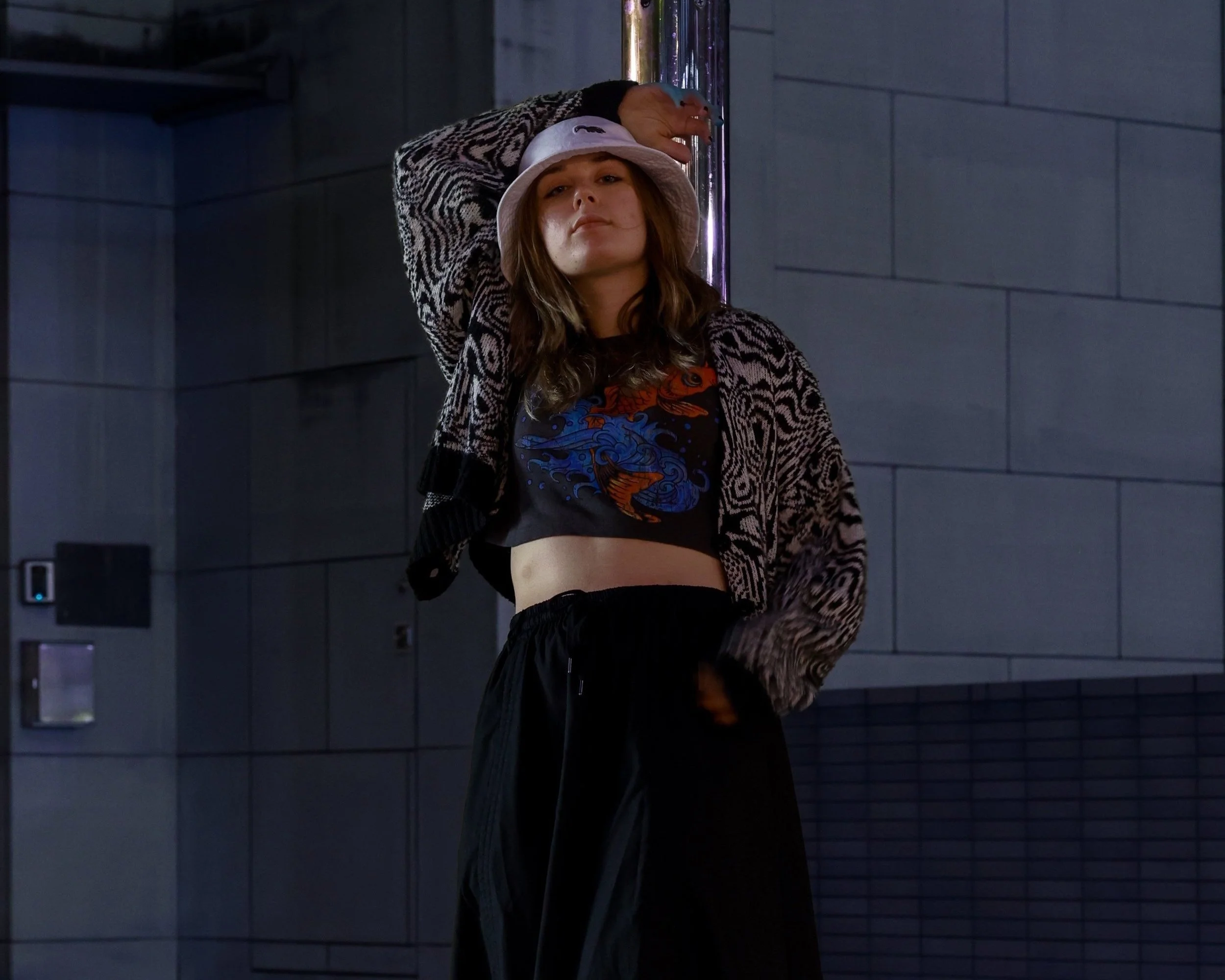 A young woman standing against a wall, wearing a white bucket hat, a patterned cardigan, a graphic t-shirt with a dragon and water design, and black pants. She has one arm resting on her head and looks confident.