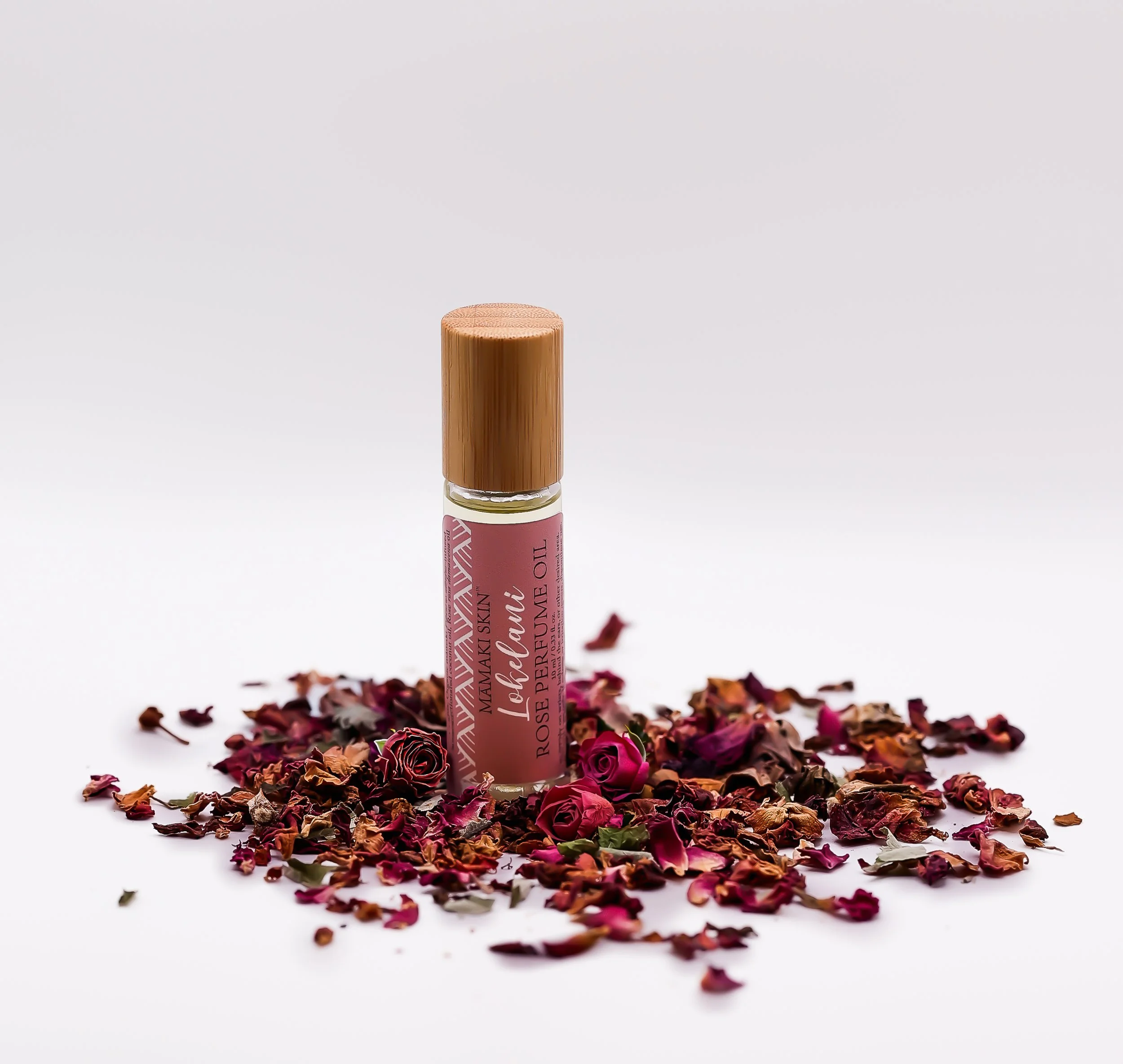 A small bottle of rose-scented perfume with a pink label, surrounded by scattered dried rose petals on a white background.