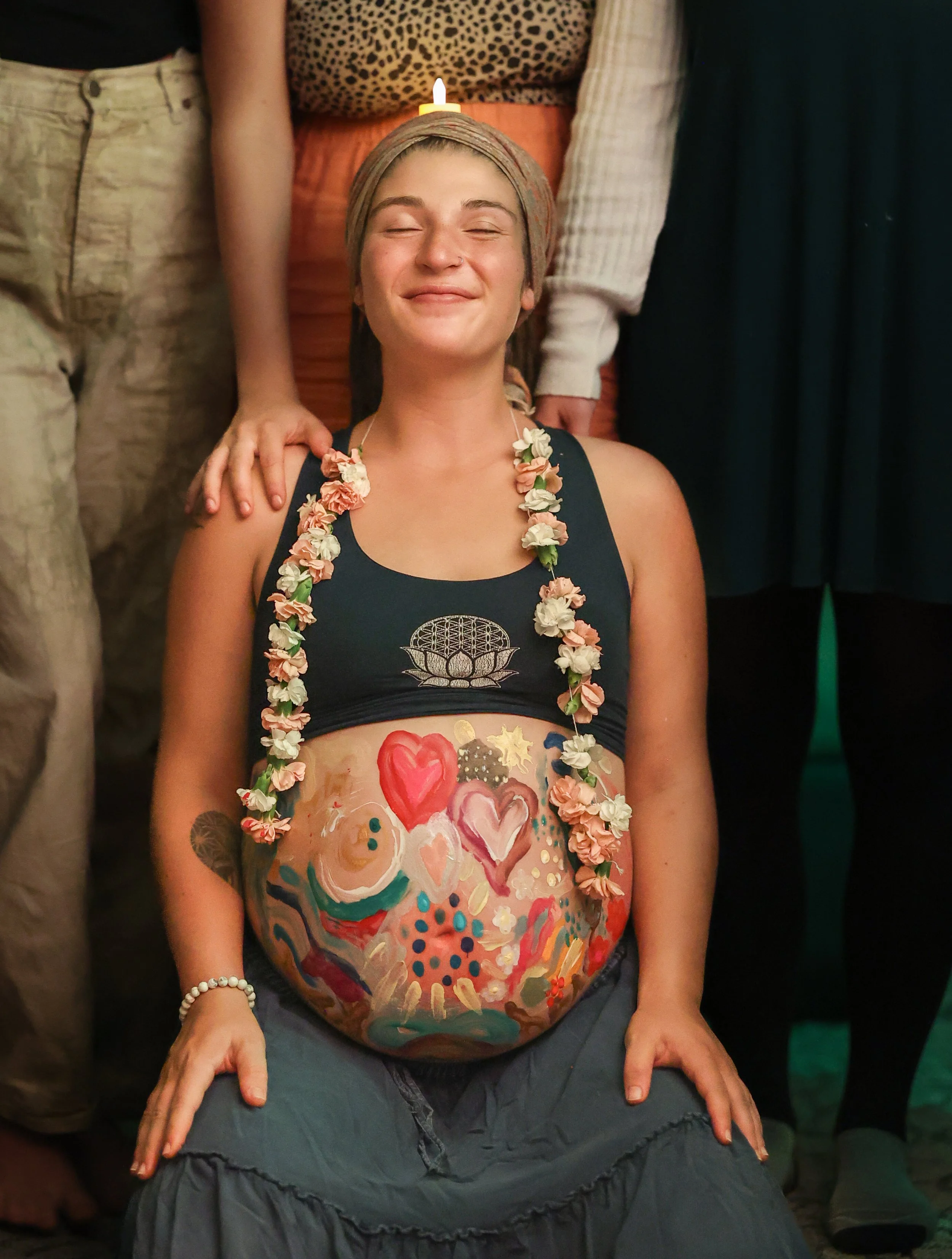 A woman with a headwrap and floral necklace smiling with her eyes closed, surrounded by people at a gathering.