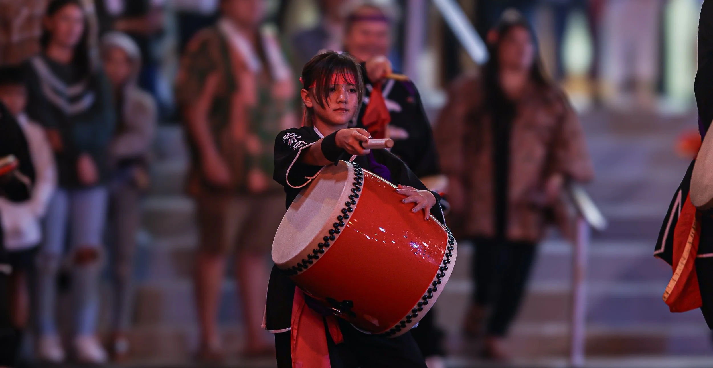 A young girl playing a large red drum during a cultural event, surrounded by other people in traditional attire in a semi-outdoor setting.