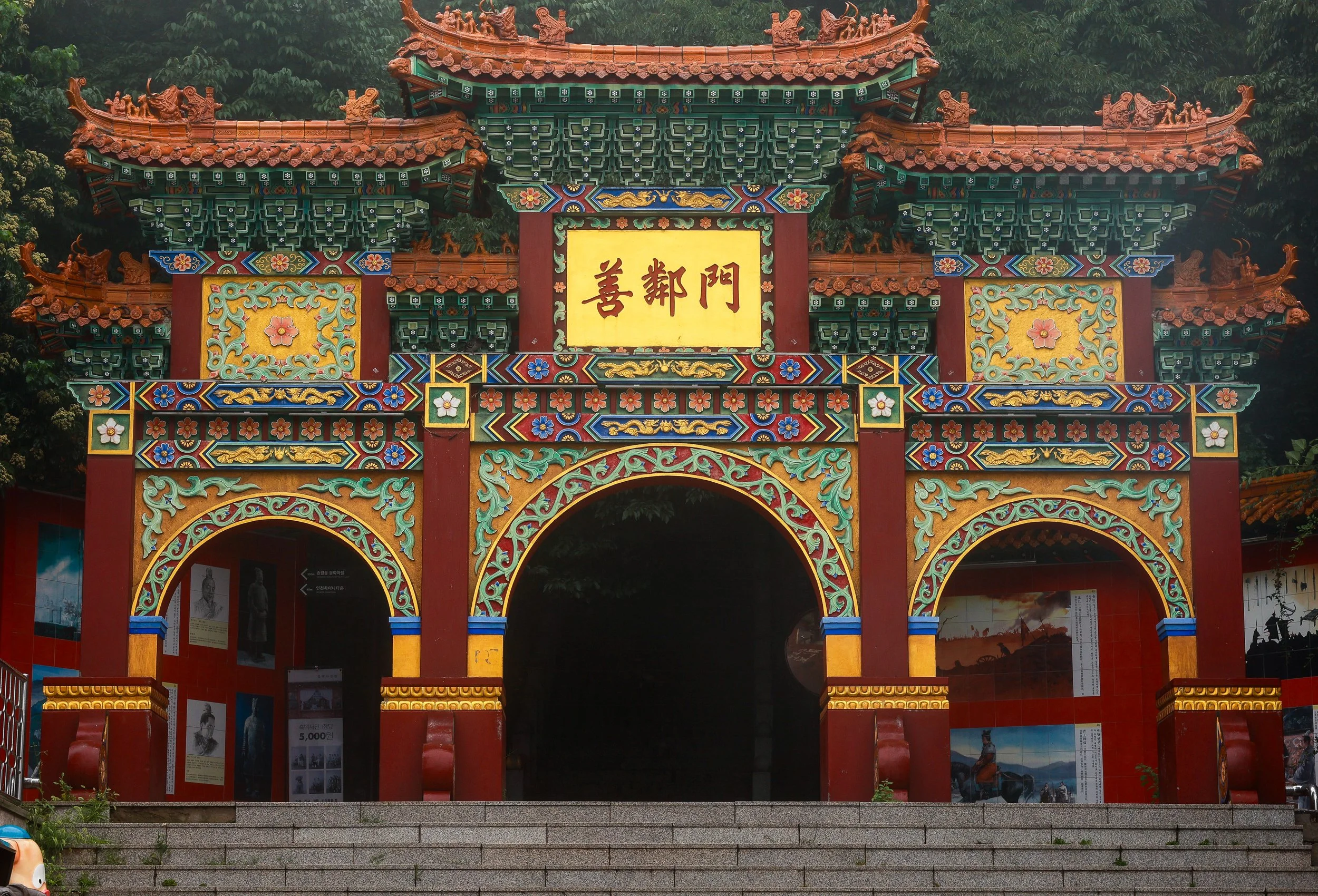 Ornate traditional Chinese temple gate with colorful decorative details and Chinese characters on a yellow sign, surrounded by greenery.