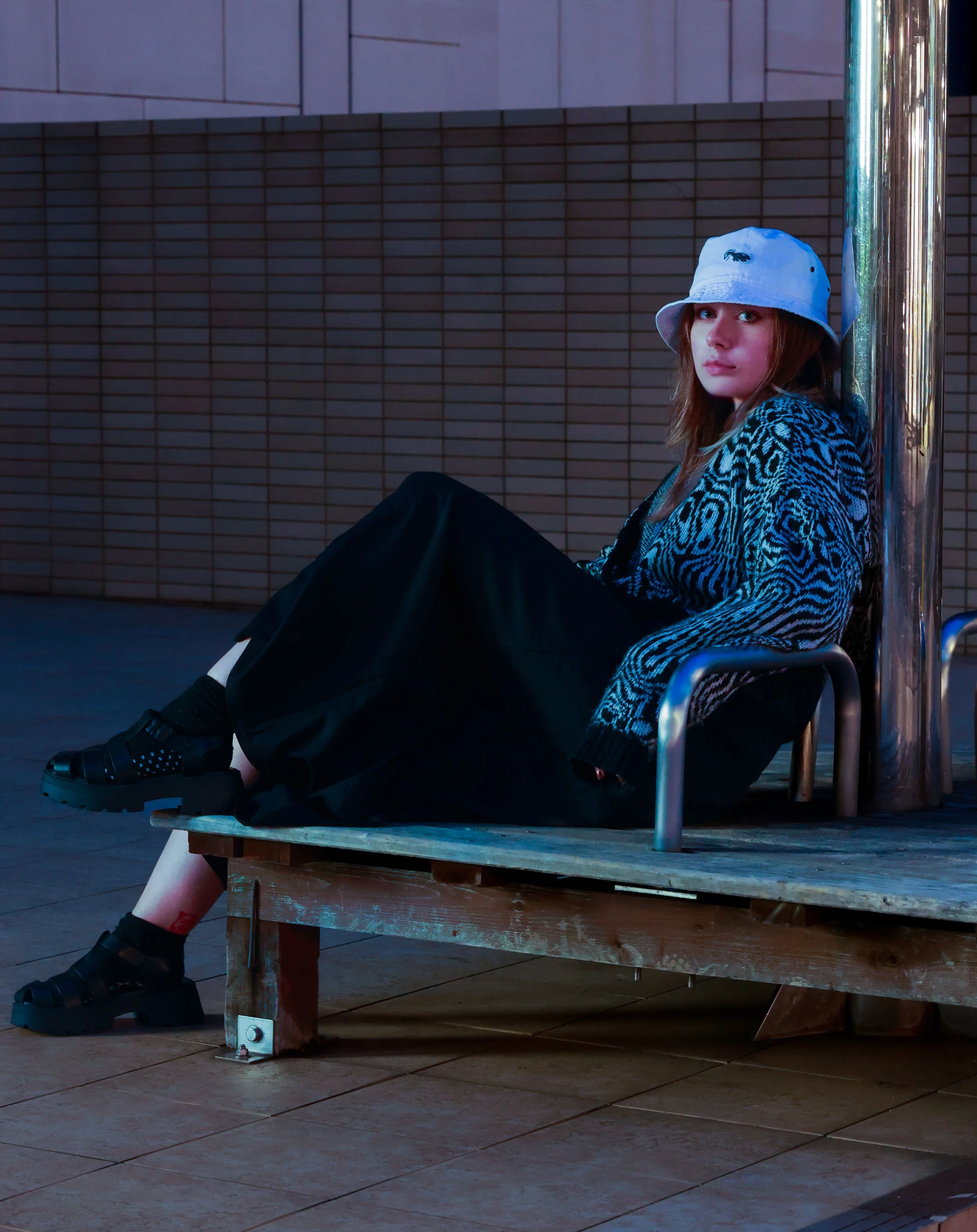 A young woman with red hair wearing a white bucket hat and black platform sandals sitting on a wooden platform against a tiled wall and metallic pole.