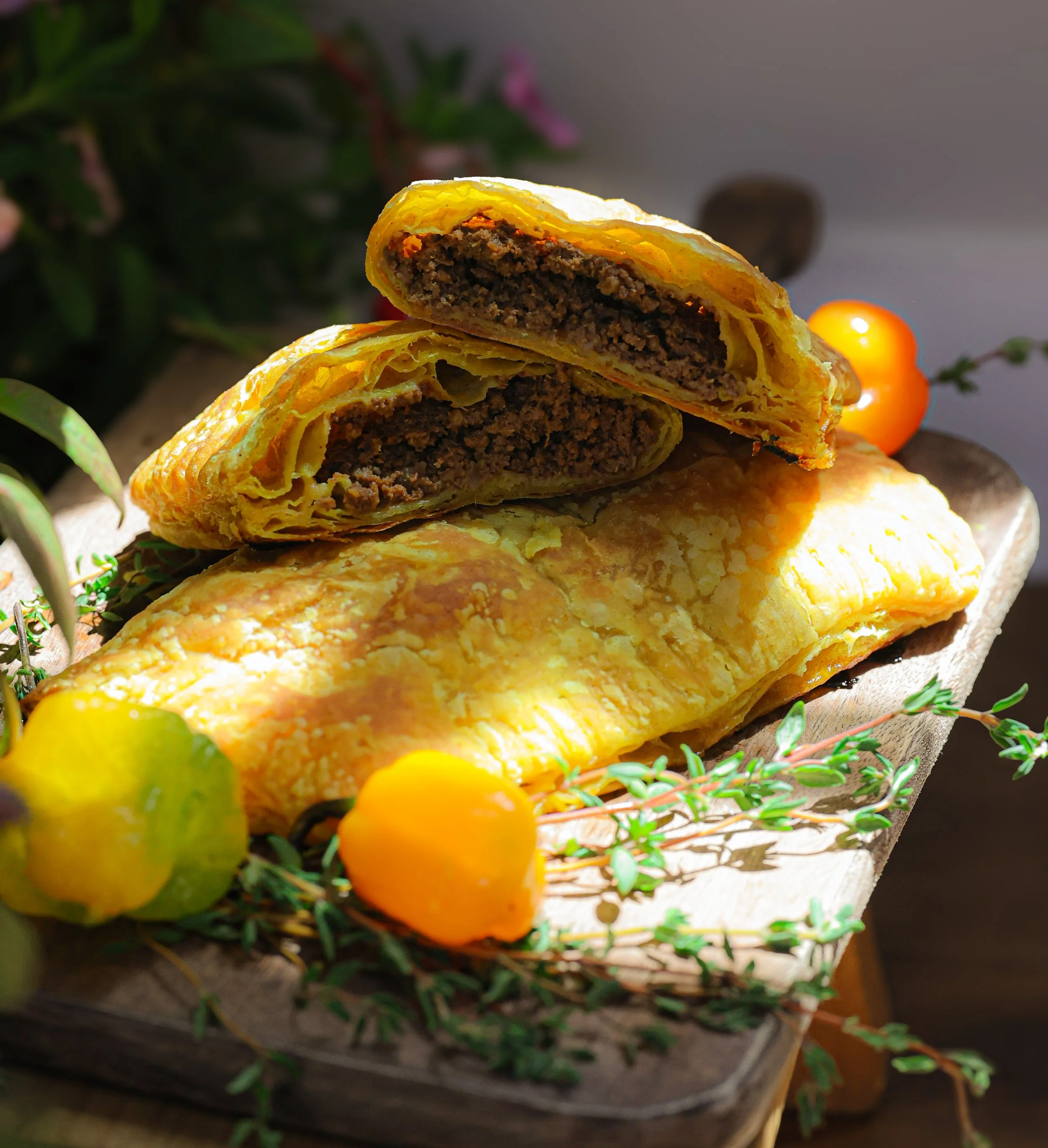 Close-up of a sliced beef and cheese pastry with a golden crust, resting on a woven basket, decorated with green leaves and small yellow and orange berries in the background.