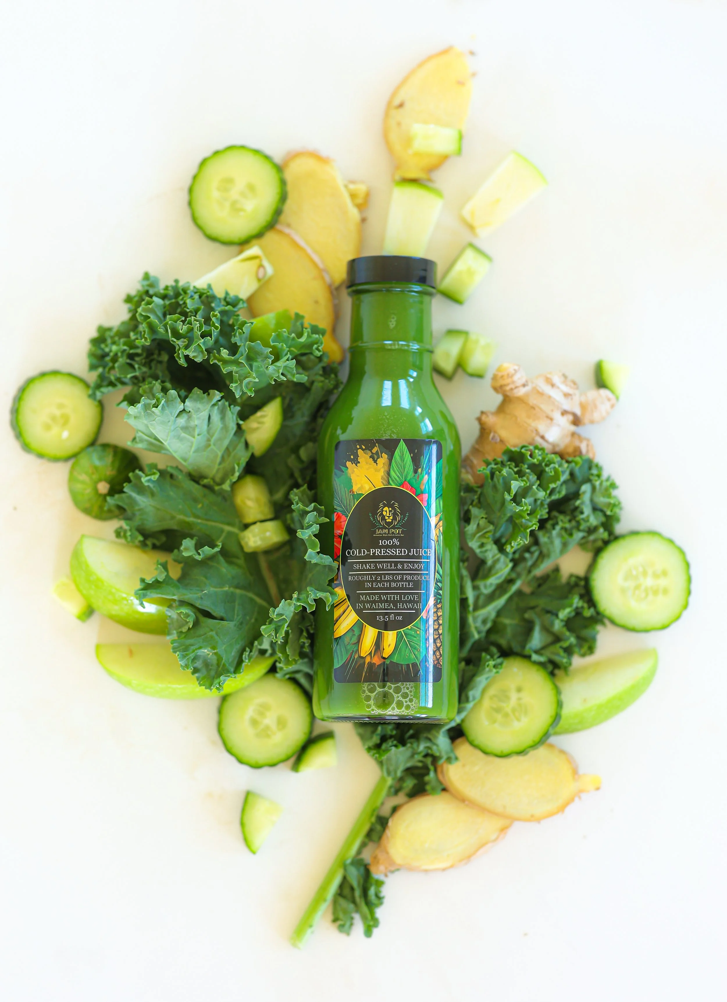 A green bottle of cold-pressed juice with tropical fruit and vegetable slices surrounding it, including cucumber, ginger, and lemon, on a white background.