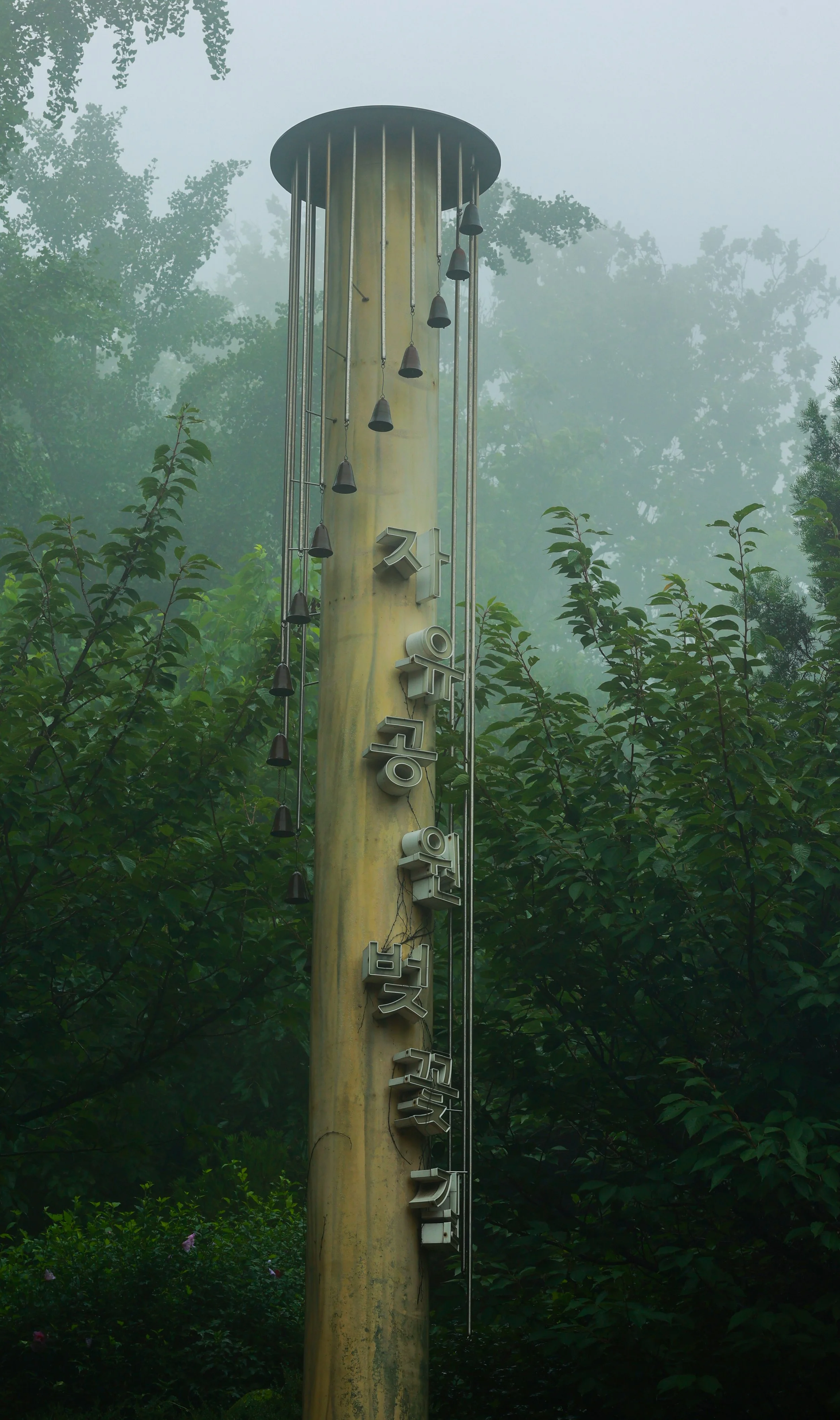 A tall yellow utility pole surrounded by green trees and foliage in foggy weather, with metal signs and hanging bells attached.