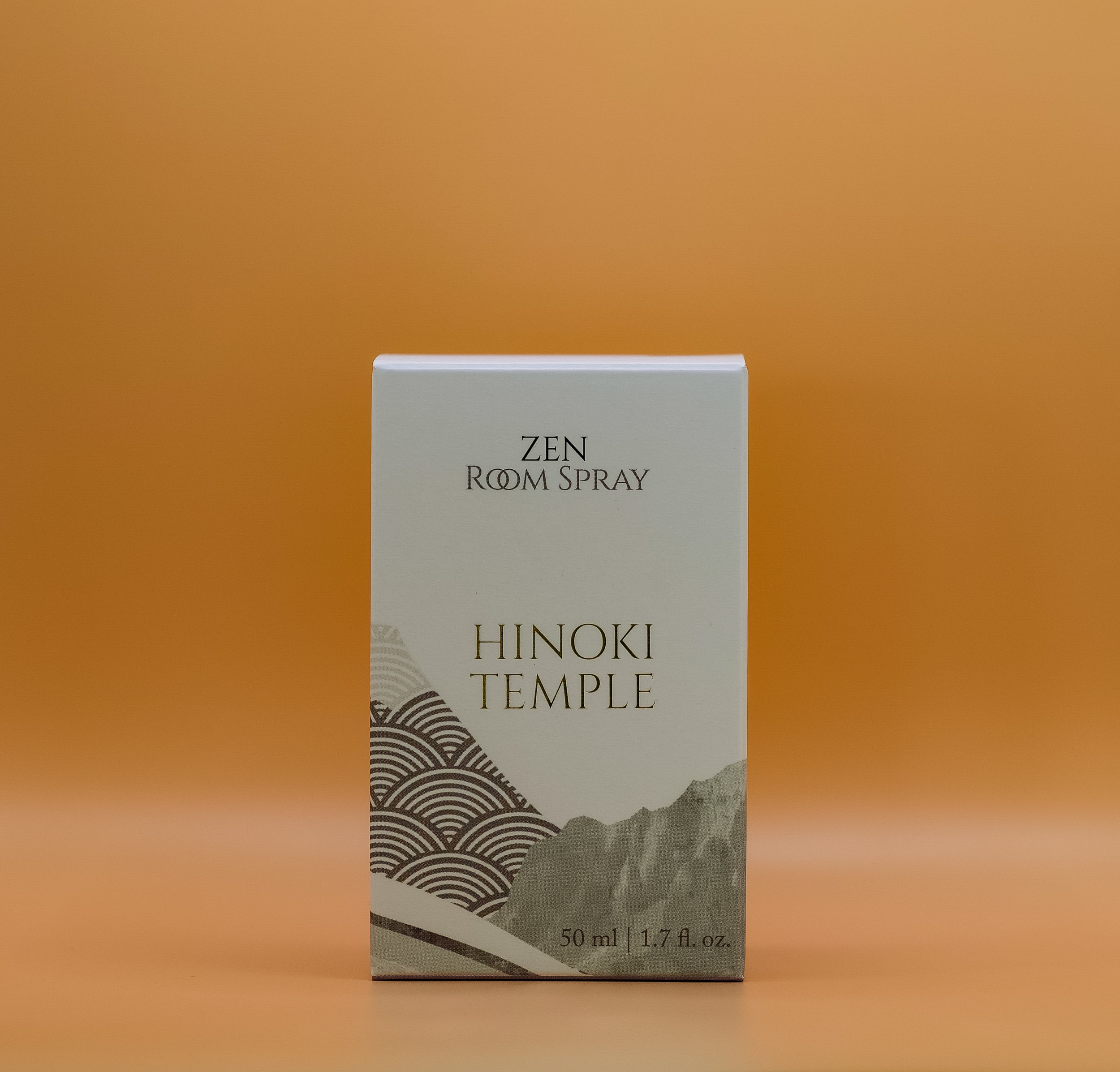 A white box of Zen Room Spray with a partly visible label featuring black text and wave-like patterns at the bottom, positioned against a plain orange wall background.