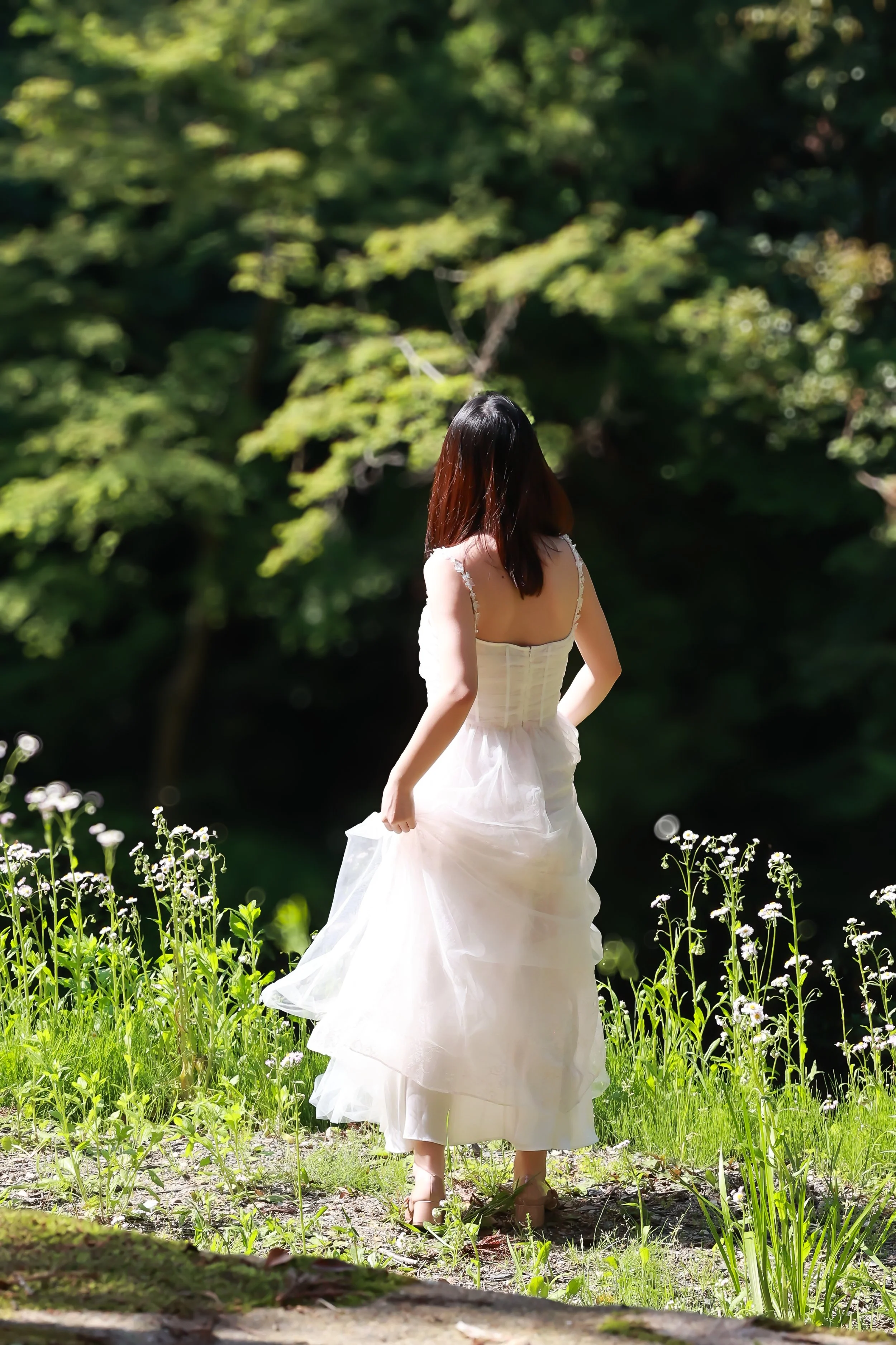 A woman in a white dress standing outdoors in a garden with green trees and flowers.