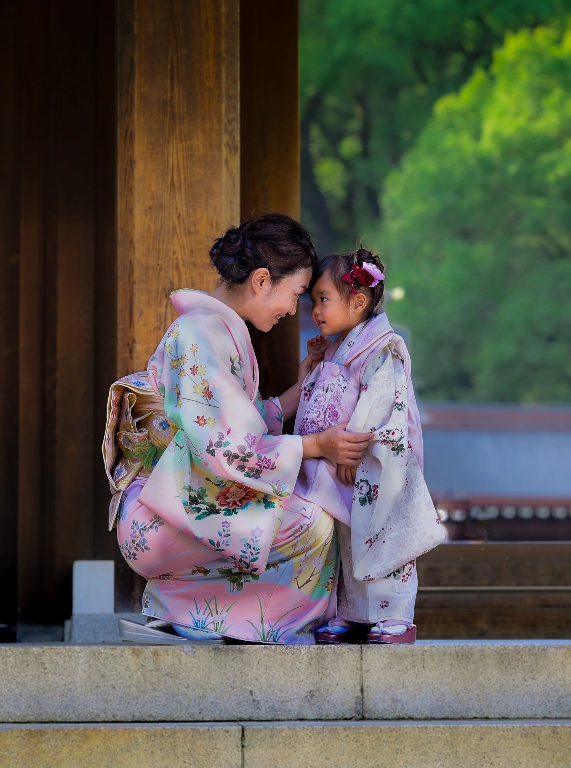 A woman and a girl dressed in traditional Japanese kimonos sharing an intimate moment on the steps of a shrine or temple, with green trees in the background.
