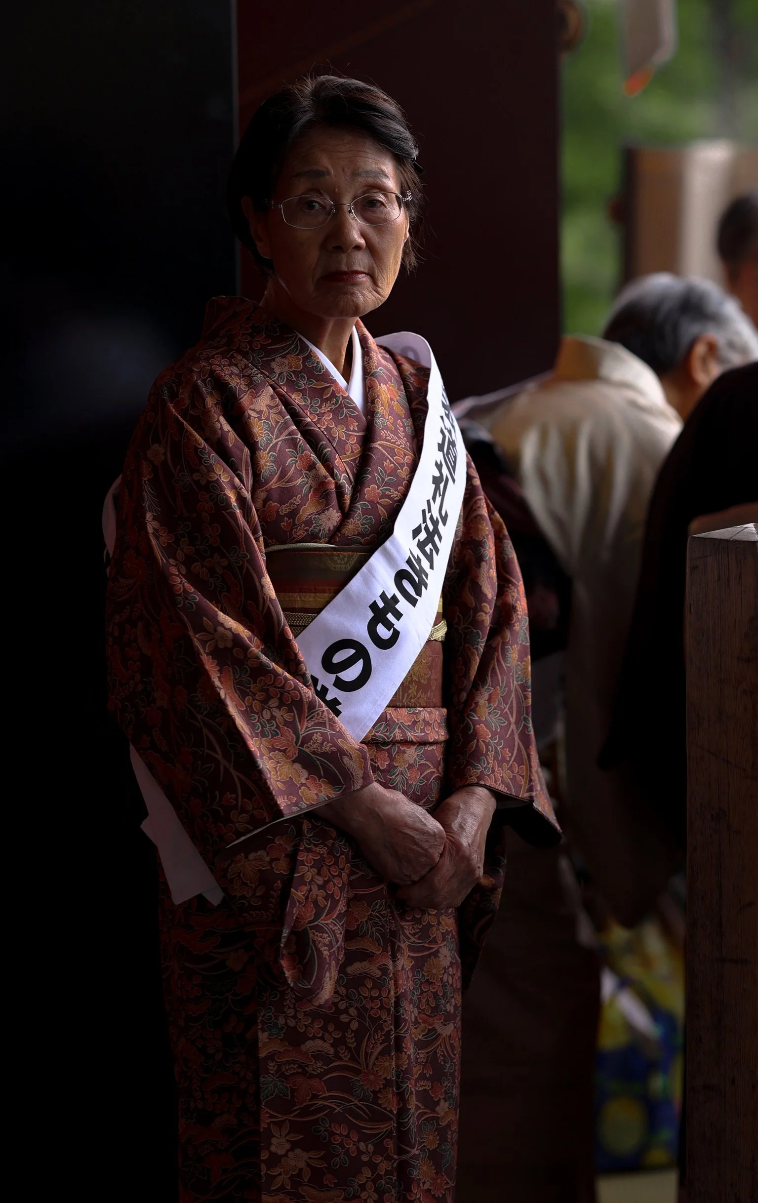 An elderly Asian woman in traditional kimono with glasses and a sash, standing with her hands clasped in front of her at a formal event.