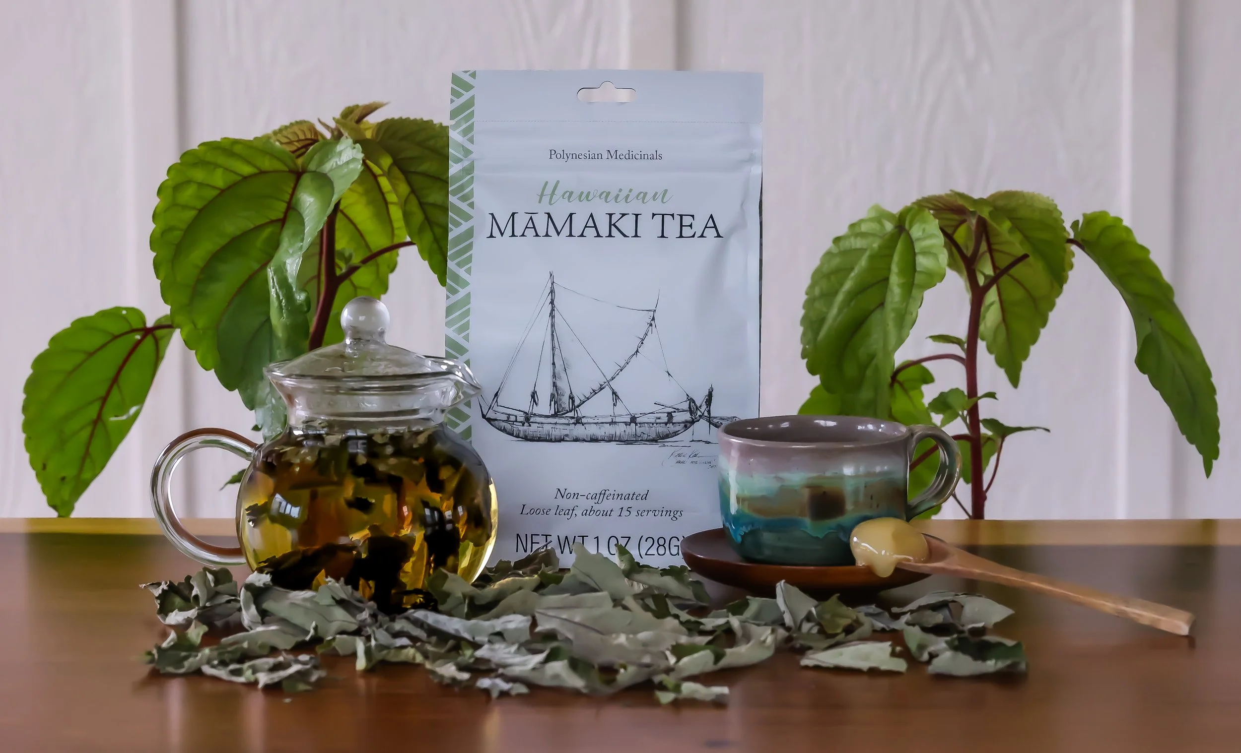 A package of Hawaiian Māmaki tea surrounded by green leafy plants, a glass teapot with loose tea leaves inside, a colorful cup filled with tea, a wooden spoon with honey, and some dried leaves on a wooden surface.