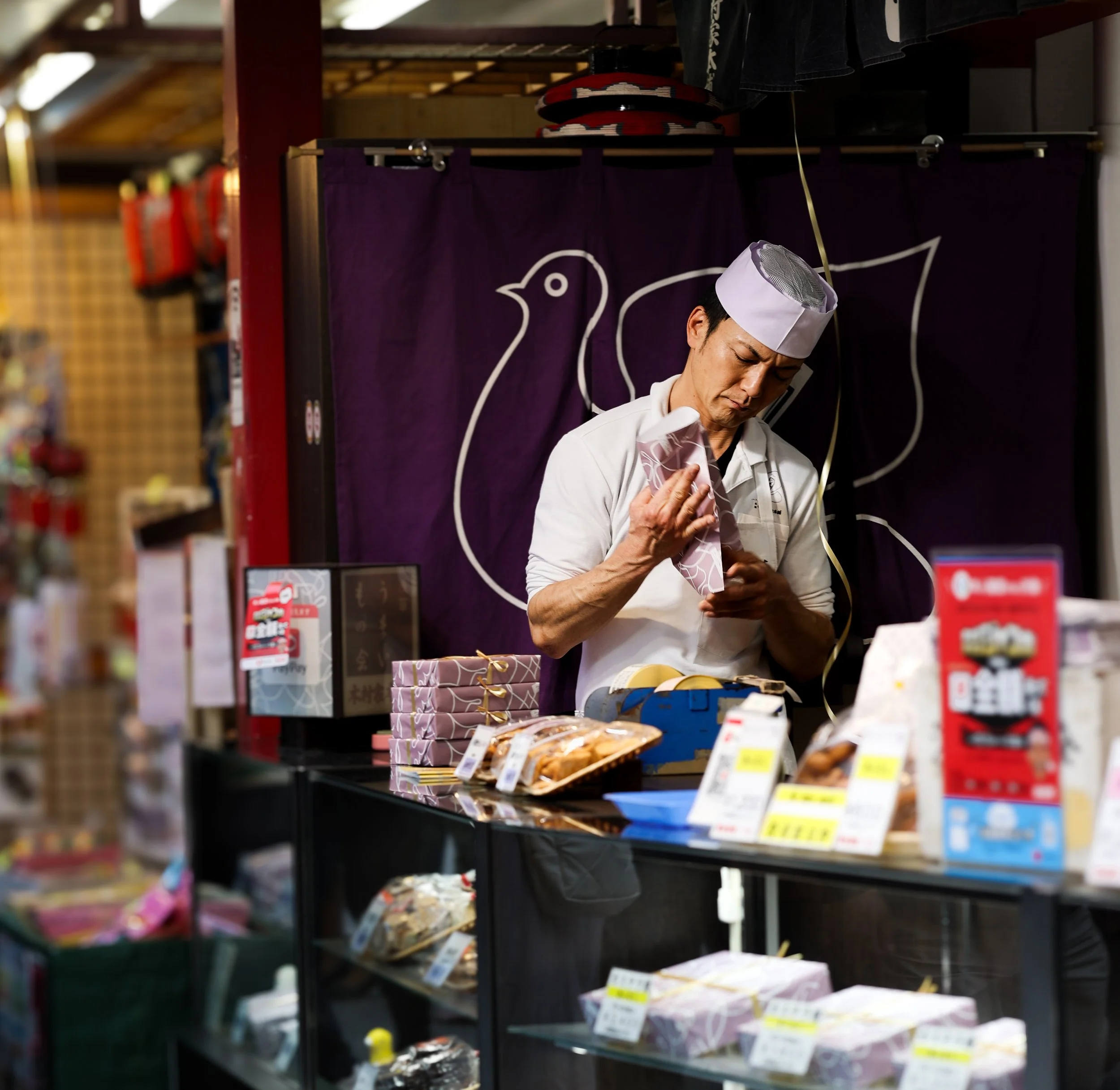 A person wearing a white chef coat and a chef hat is shopping at a grocery store, examining a package. There are various packaged food items on the shelves.