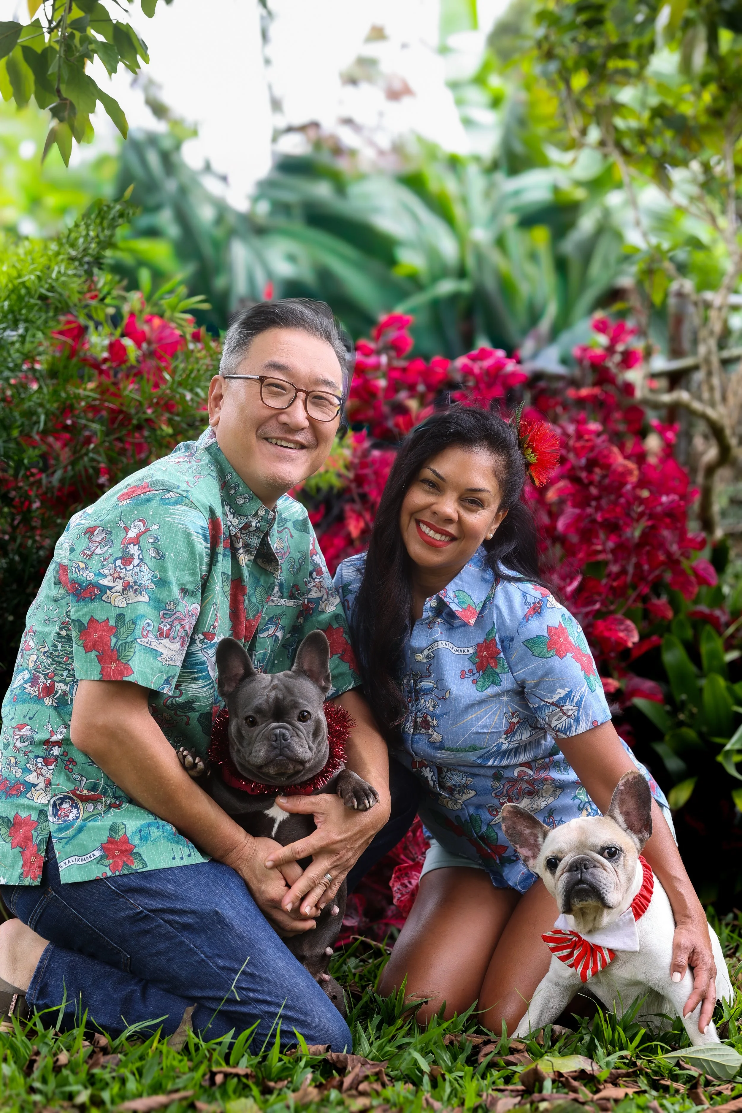 A smiling man and woman wearing tropical patterned shirts posing with two French Bulldog dogs in a garden with red and green foliage, one dog with a red collar and bow tie, the other with a red tinsel collar.