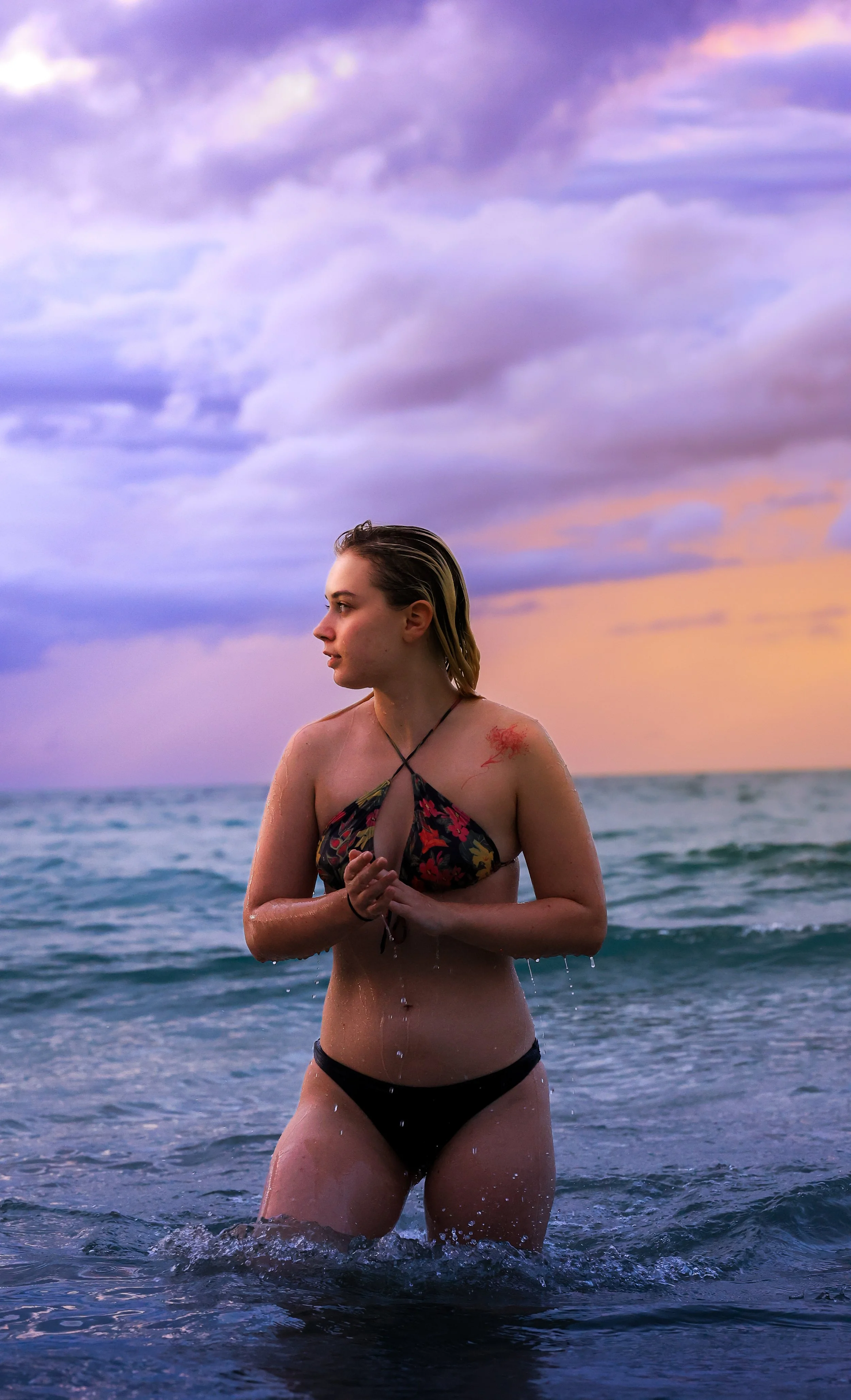 A young woman standing in the ocean during sunset, wearing a floral bikini top and black bikini bottoms, with her hands clasped in front of her, and a red tattoo on her shoulder.