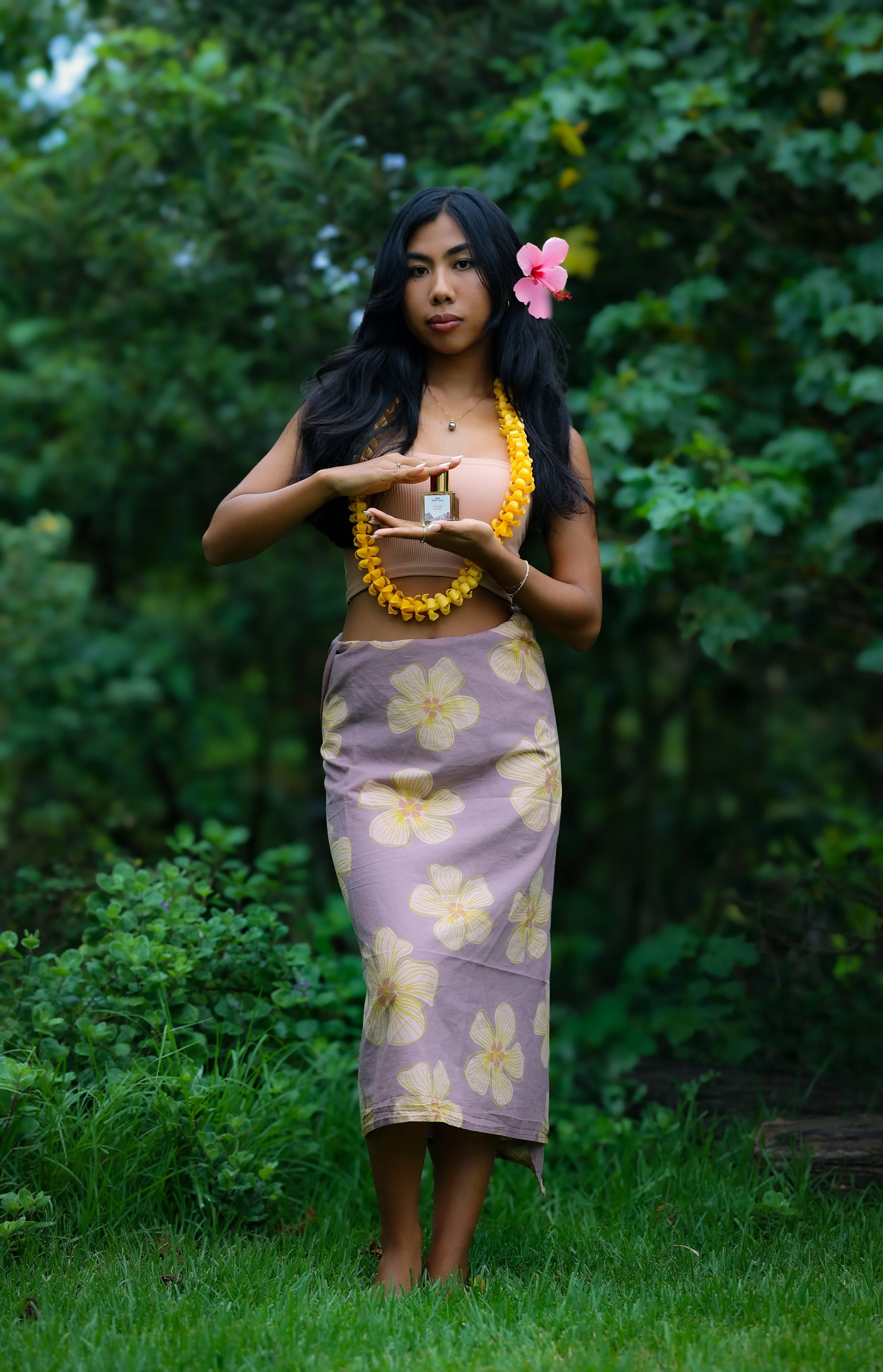 A woman standing outdoors in a lush garden, wearing a vintage floral skirt, a top, and a yellow lei, with a pink hibiscus flower in her hair, holding a perfume bottle in her hands.