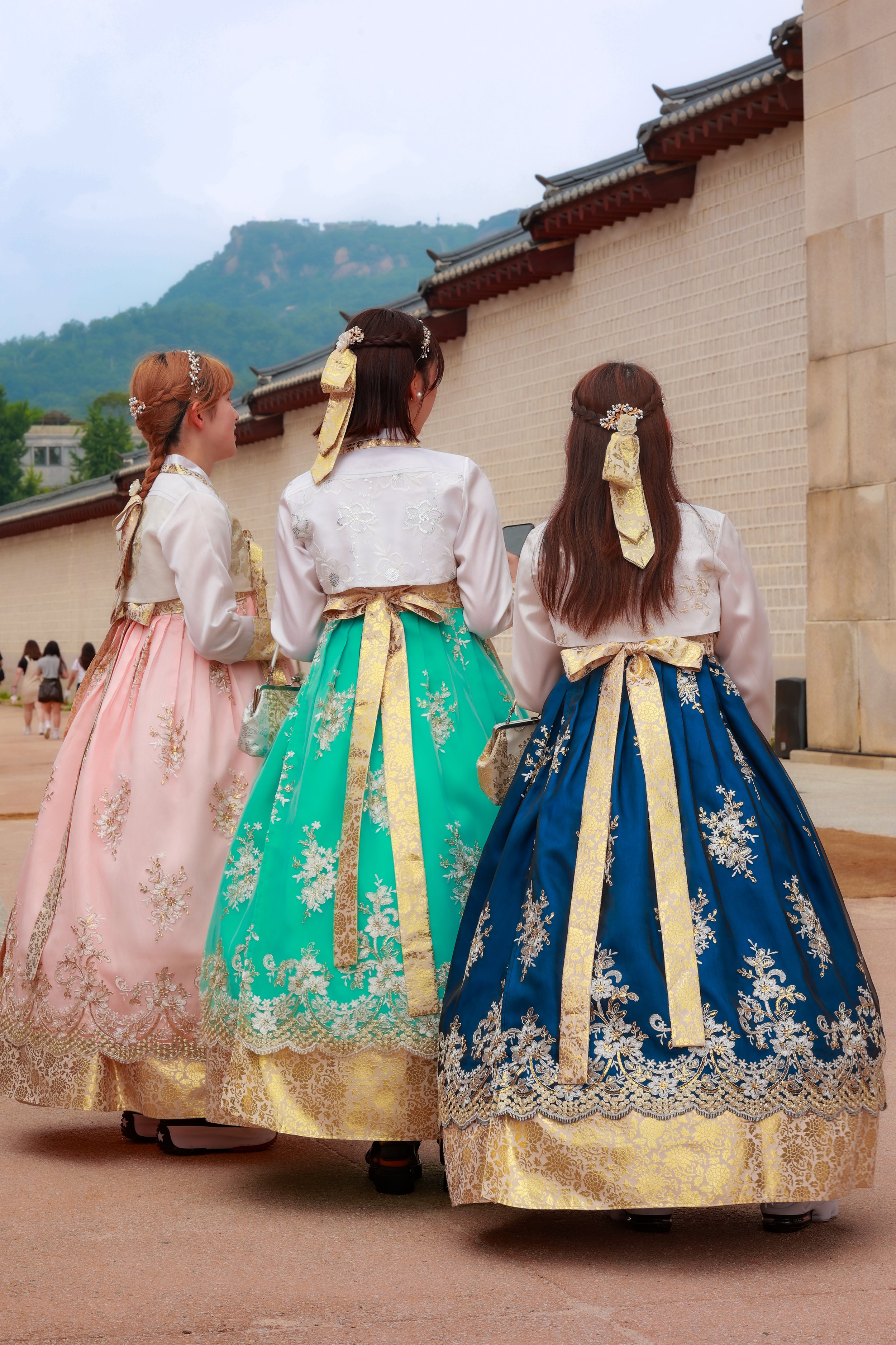 Three women dressed in traditional Korean hanbok, seen from the back, walking outdoors near a wall with a mountain in the background.
