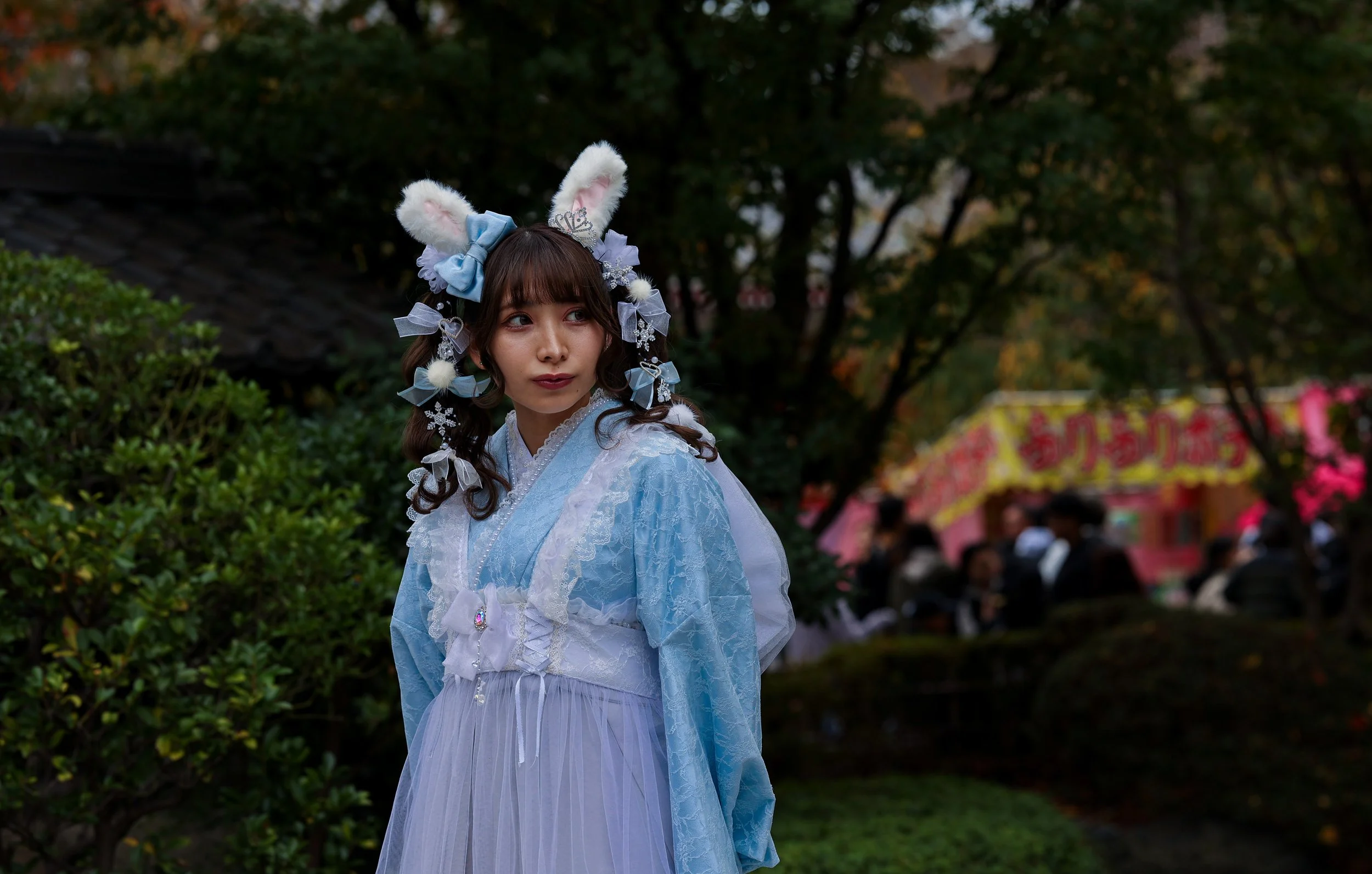 A woman dressed in a light blue fairy-tale inspired dress with rabbit ears, bows, and snowflake accessories, standing outdoors with trees and a marketplace or festival in the background.