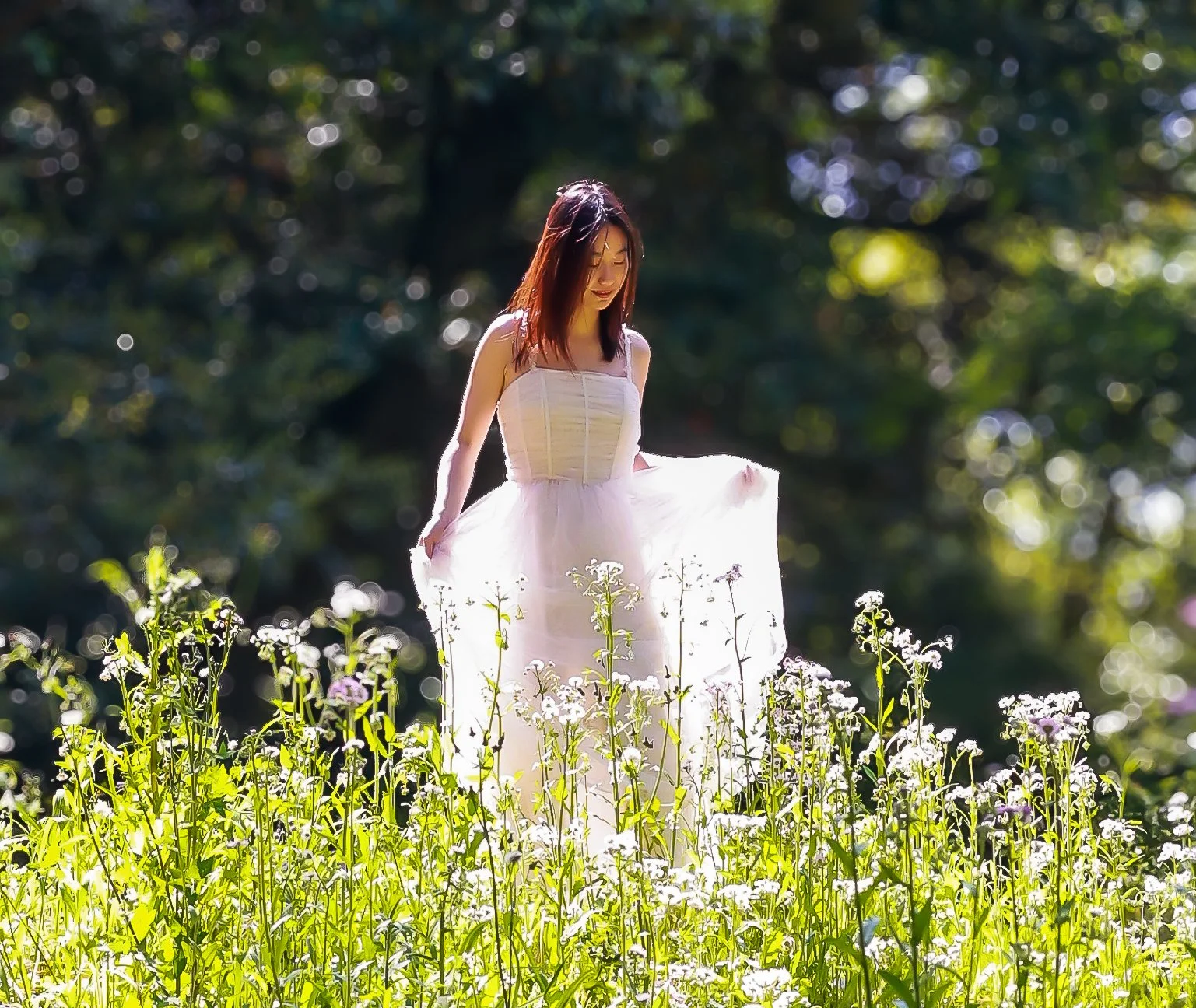 A woman in a white dress stands in a field of flowers, with a blurred green forest background.