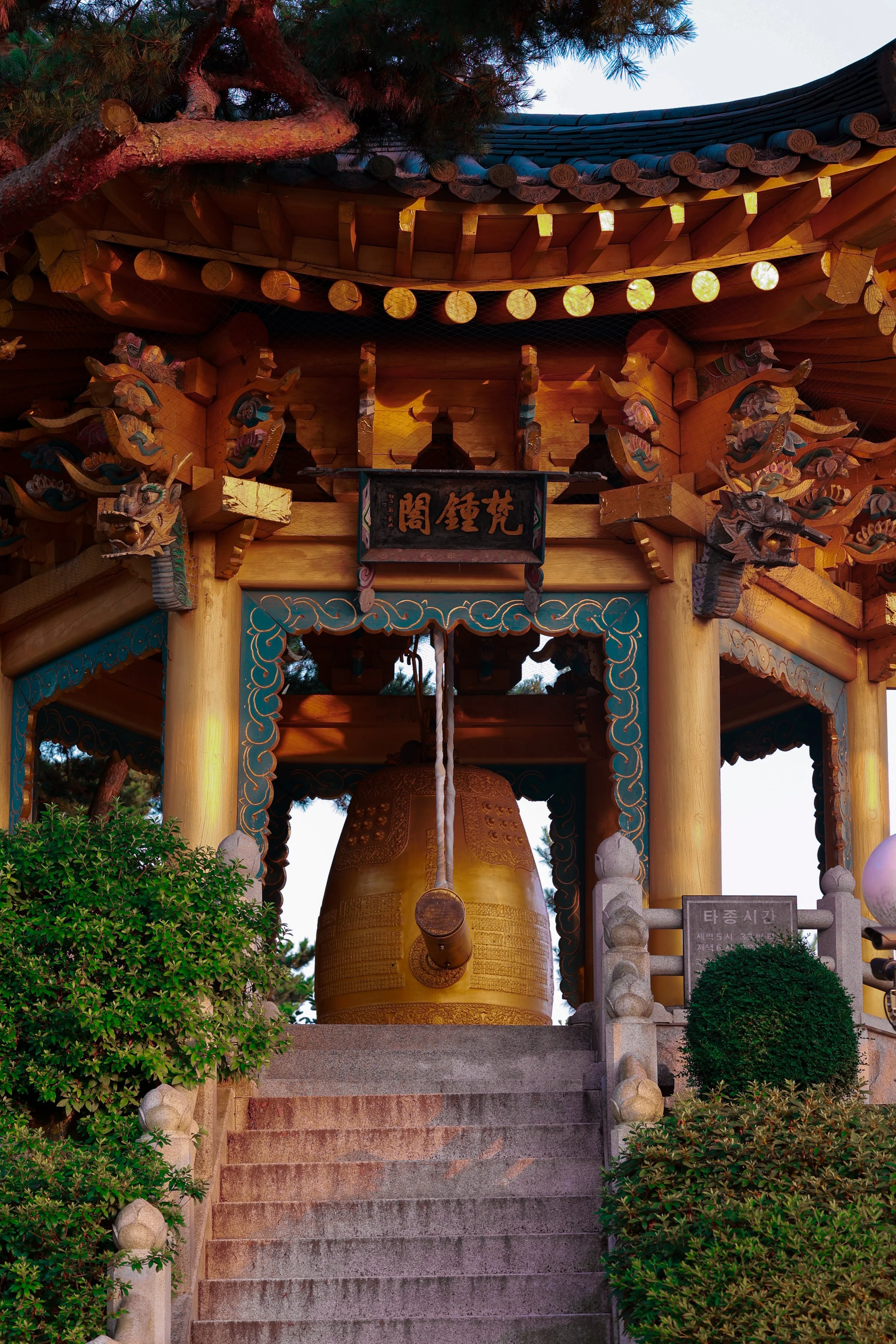 Traditional Korean temple gate with a large golden bell hanging beneath, intricate wooden carvings, tiled roof, and lush greenery flanking the entrance.
