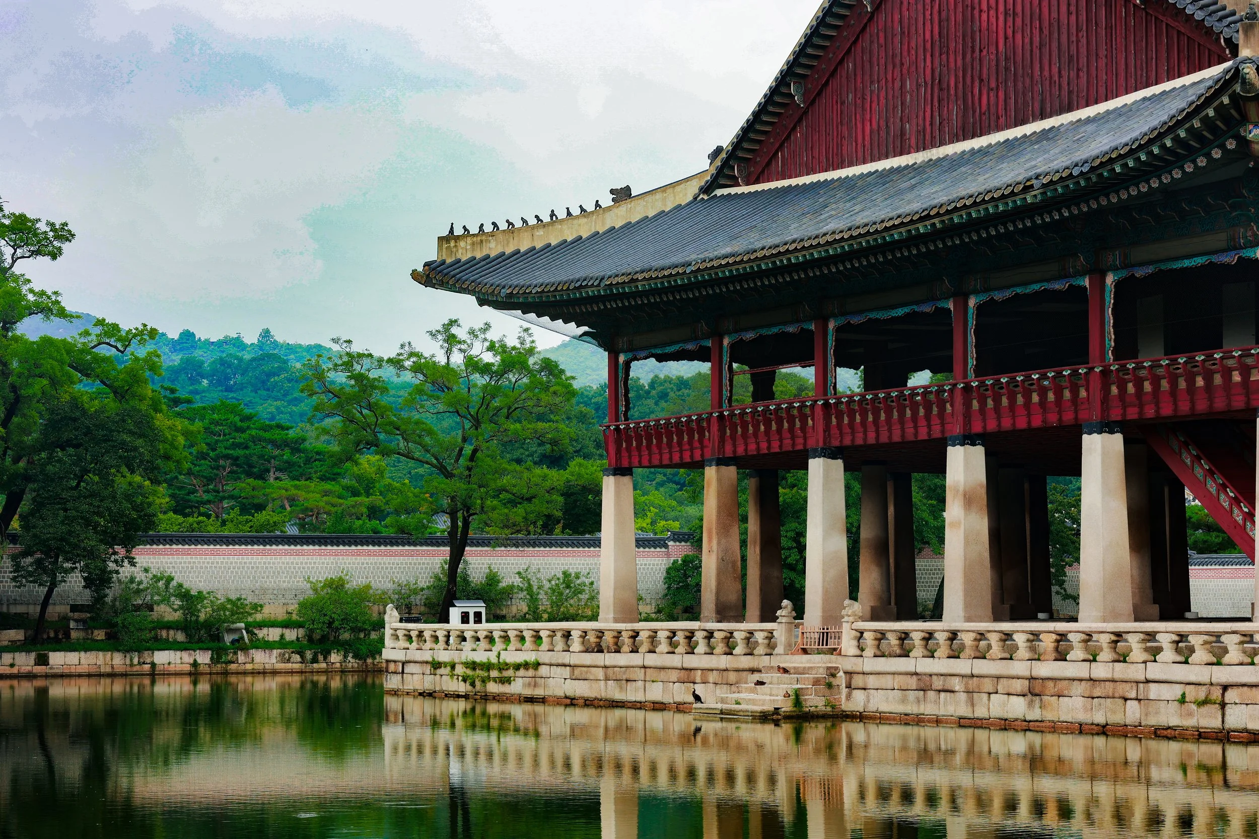 A traditional Asian pavilion with a curved roof, red and dark tiles, situated next to a water body with trees and a stone wall in the background.