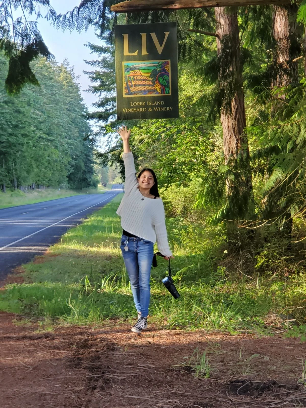 A woman standing by a roadside in a forested area, smiling and holding a camera in one hand while reaching up with the other hand to point at a sign that reads 'LIV Lopez Island Vineyard & Winery'.