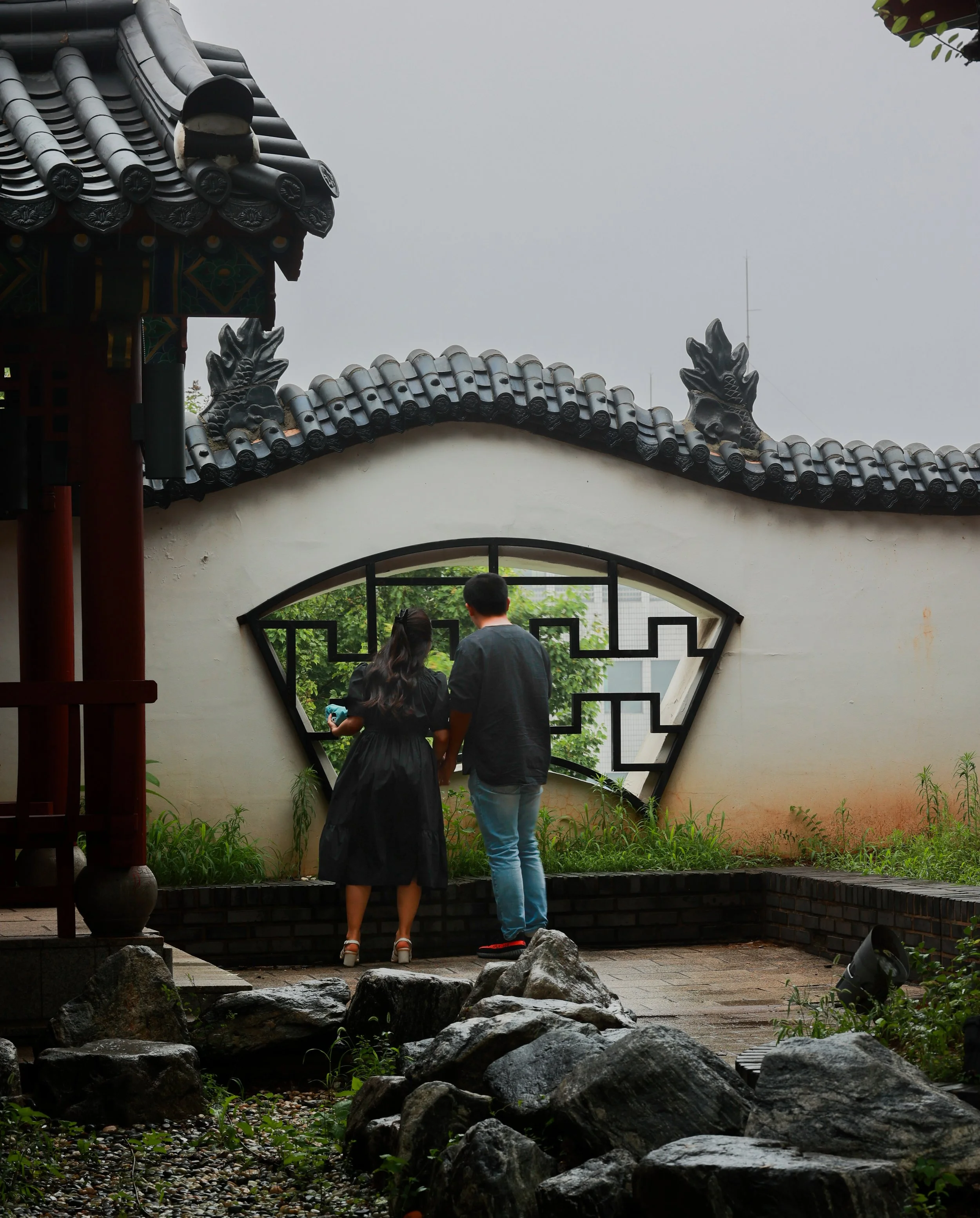 Two people, a woman in a black dress and a man in a black shirt and light blue jeans, stand together in front of a traditional Asian-style building with ornate roof decorations and a decorative window, surrounded by rocks and greenery.