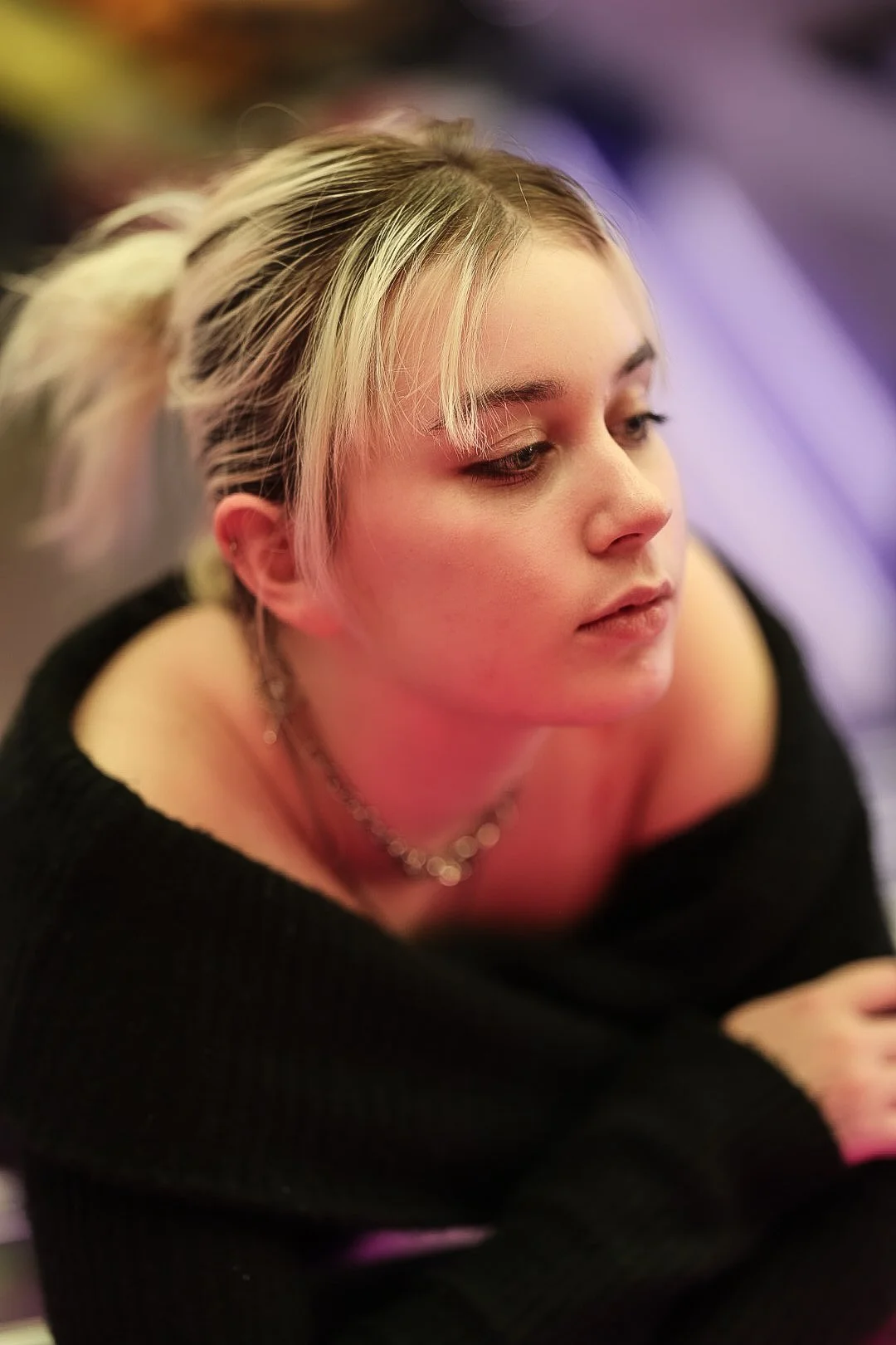 A young woman with blonde hair styled in pigtails, wearing a black off-shoulder top and a silver necklace, looking to the side in a softly lit setting.
