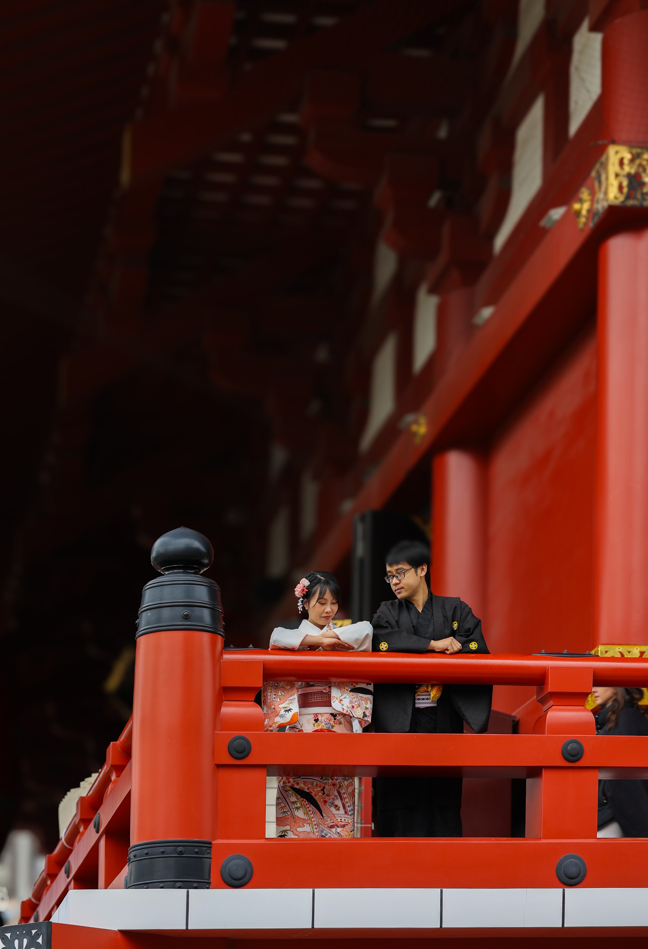 A young woman in a kimono standing on a red bridge, with a young man in a black traditional outfit, looking at her. The bridge has black lamps and red wooden railings, and the background is dark.