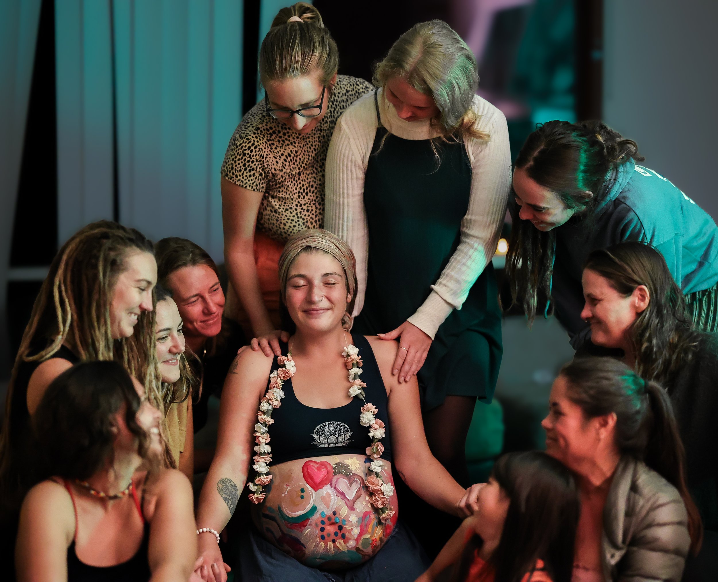 A group of people gathered closely together, smiling and engaged in a joyful moment. One woman in the center has painted hearts and other designs on her bare belly, suggesting a celebration or gathering related to personal expression or body art.