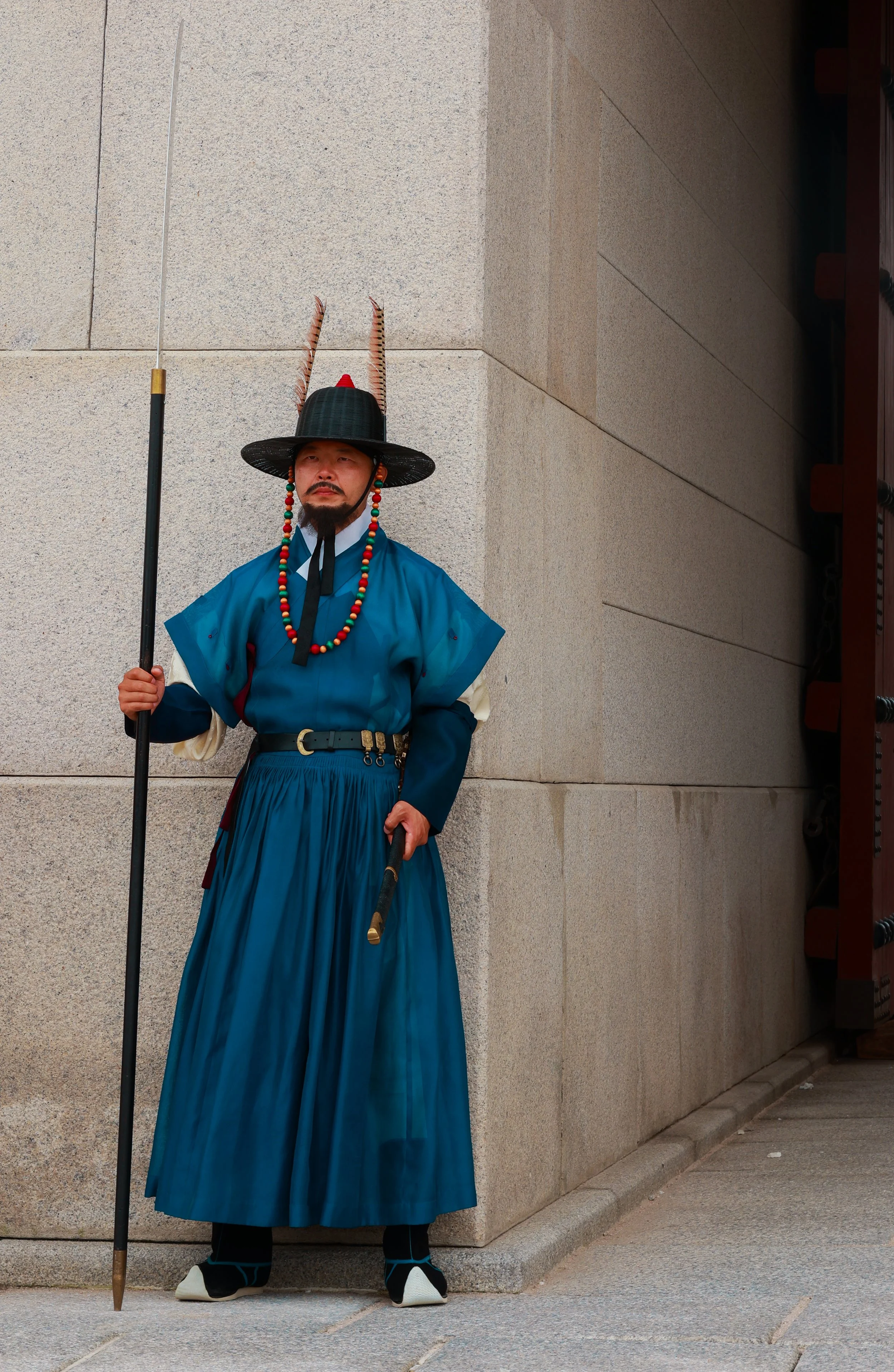 A person dressed in traditional Korean clothing, standing with a spear in front of a stone wall.