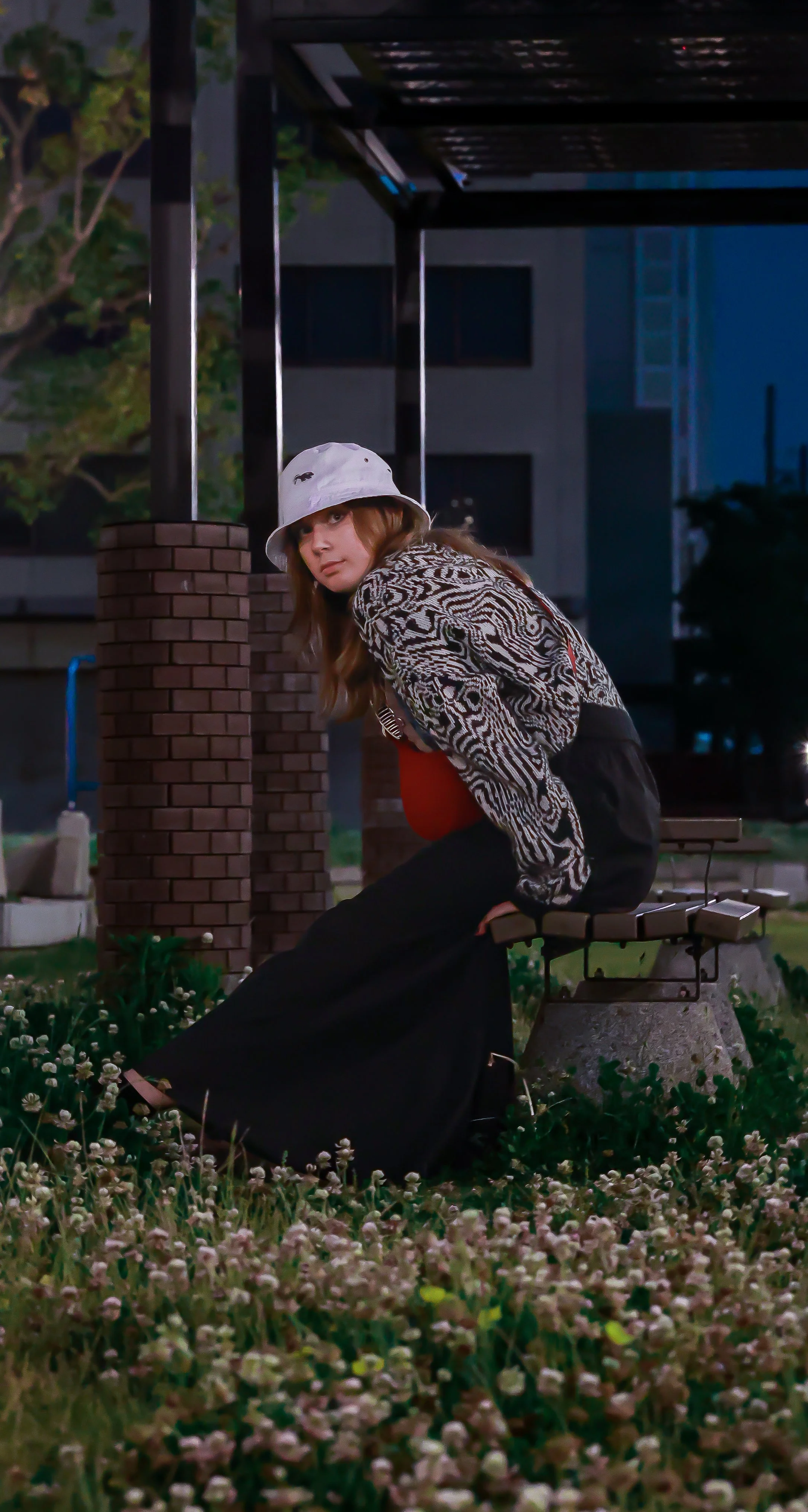 A young woman with long hair sitting on a park bench at night, wearing a patterned jacket, a white bucket hat, and black pants, surrounded by flowers with a building in the background.
