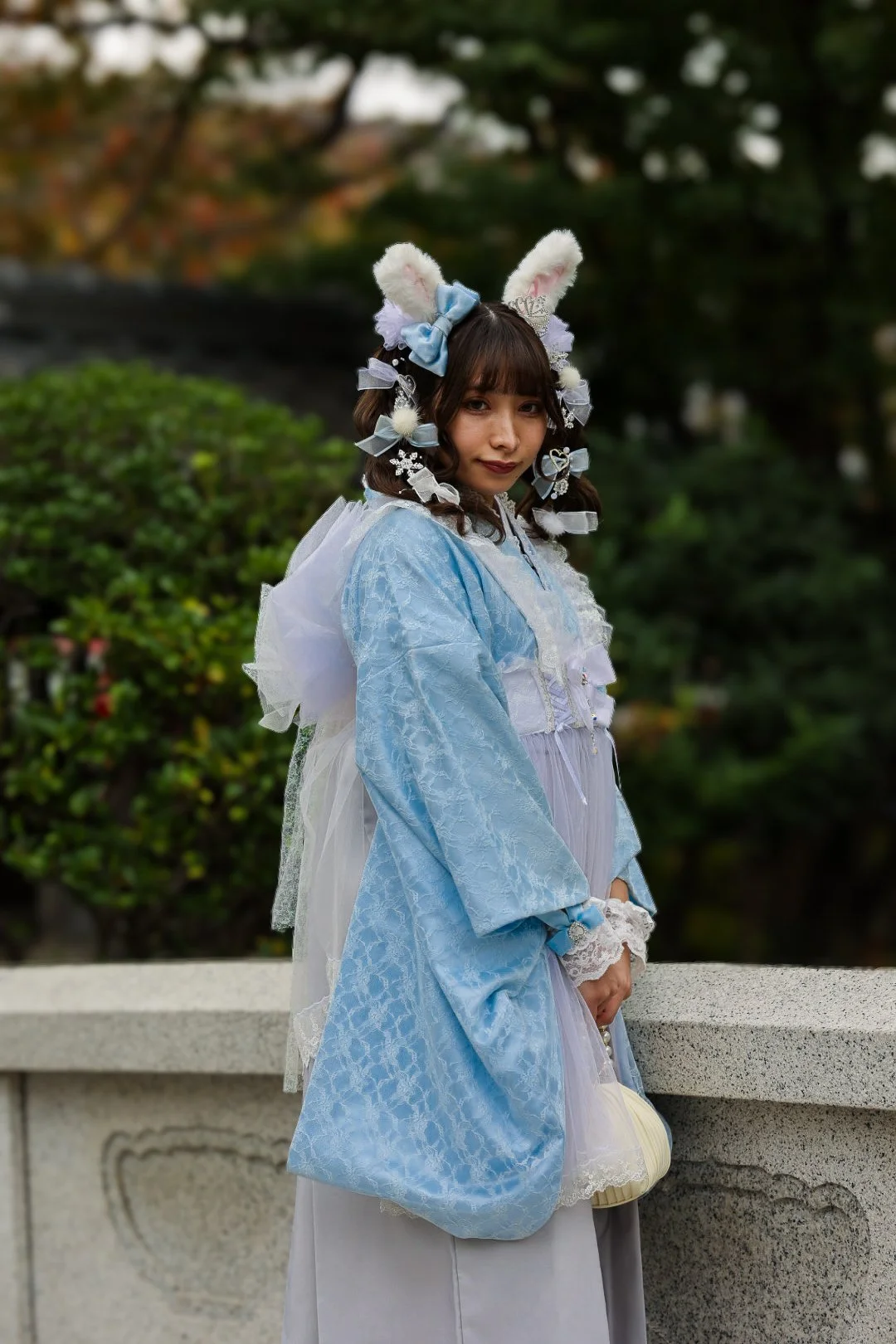 A young woman dressed in a pastel blue, lacy, and ruffled costume with white lace accents. She wears fluffy white bunny ears with pink interiors and has her hair decorated with bows, lace, and fluffy accessories. She is standing outdoors against a bl