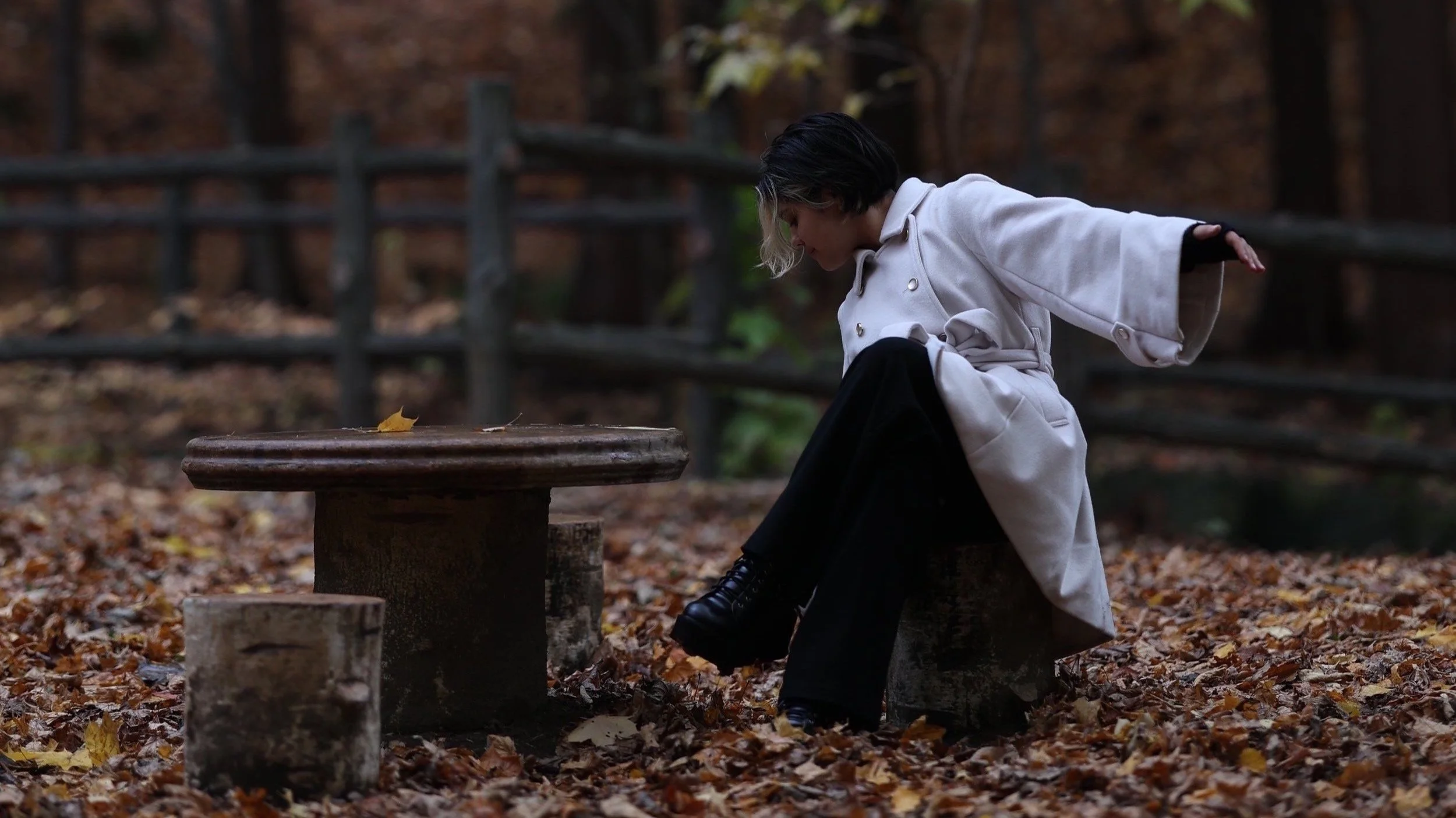A woman with short black and white hair wearing a white coat, black pants, and boots, is sitting outdoors in a wooded area with fallen autumn leaves, leaning forward with arms outstretched behind her.