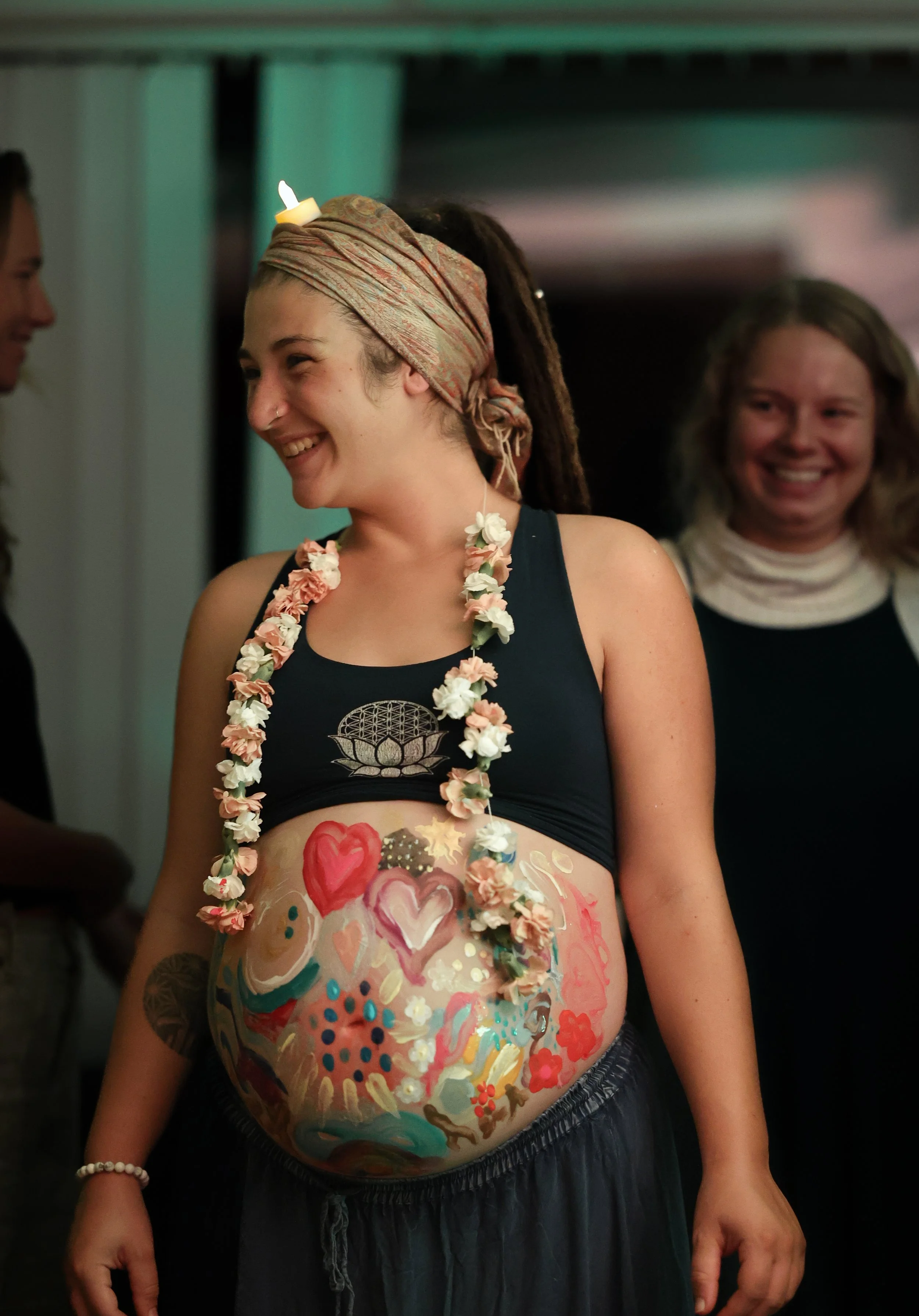 A woman with dreadlocks and a head wrap smiling, wearing a flower lei and a black crop top with a painted heart design, standing indoors with two other women in the background.
