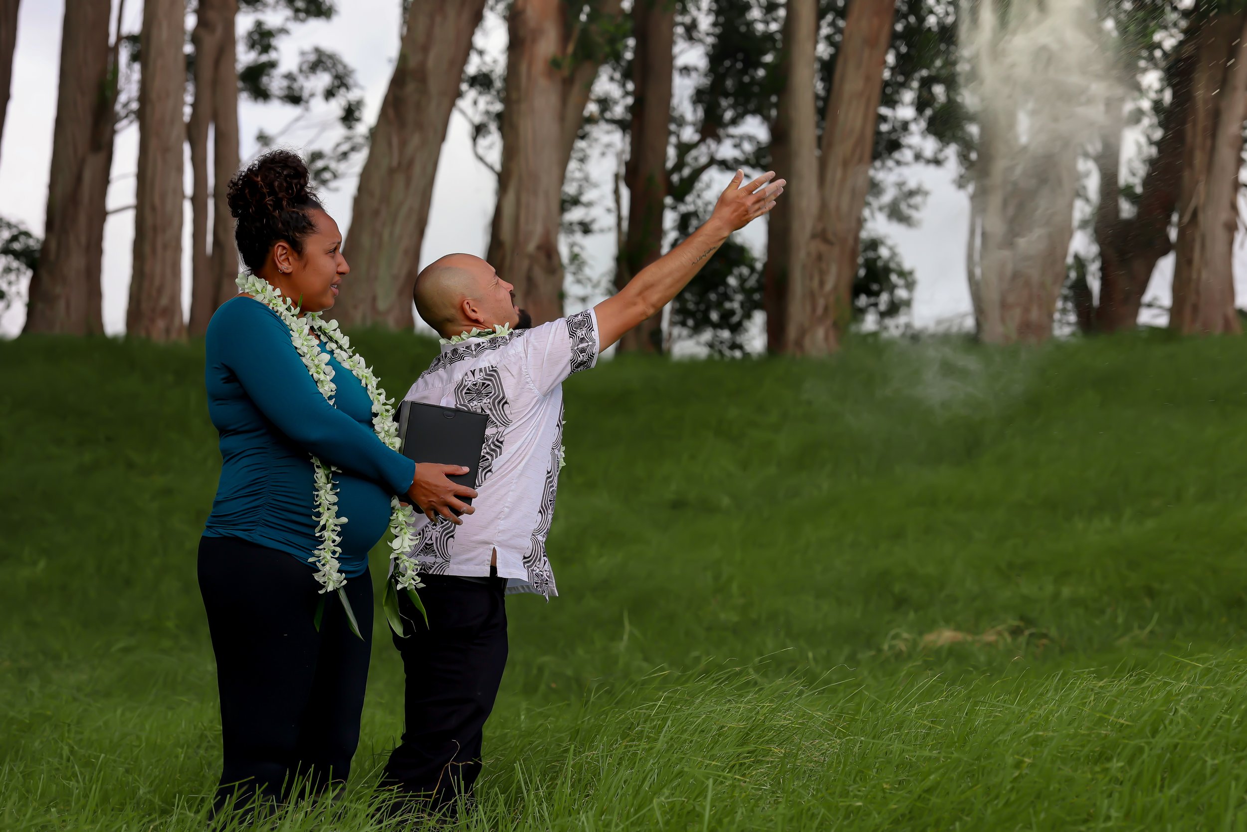 A woman and a man stand outdoors in a grassy area with tall trees in the background. The woman wears a blue outfit and a floral necklace. The man is wearing a patterned shirt and is gesturing with his hand while speaking.