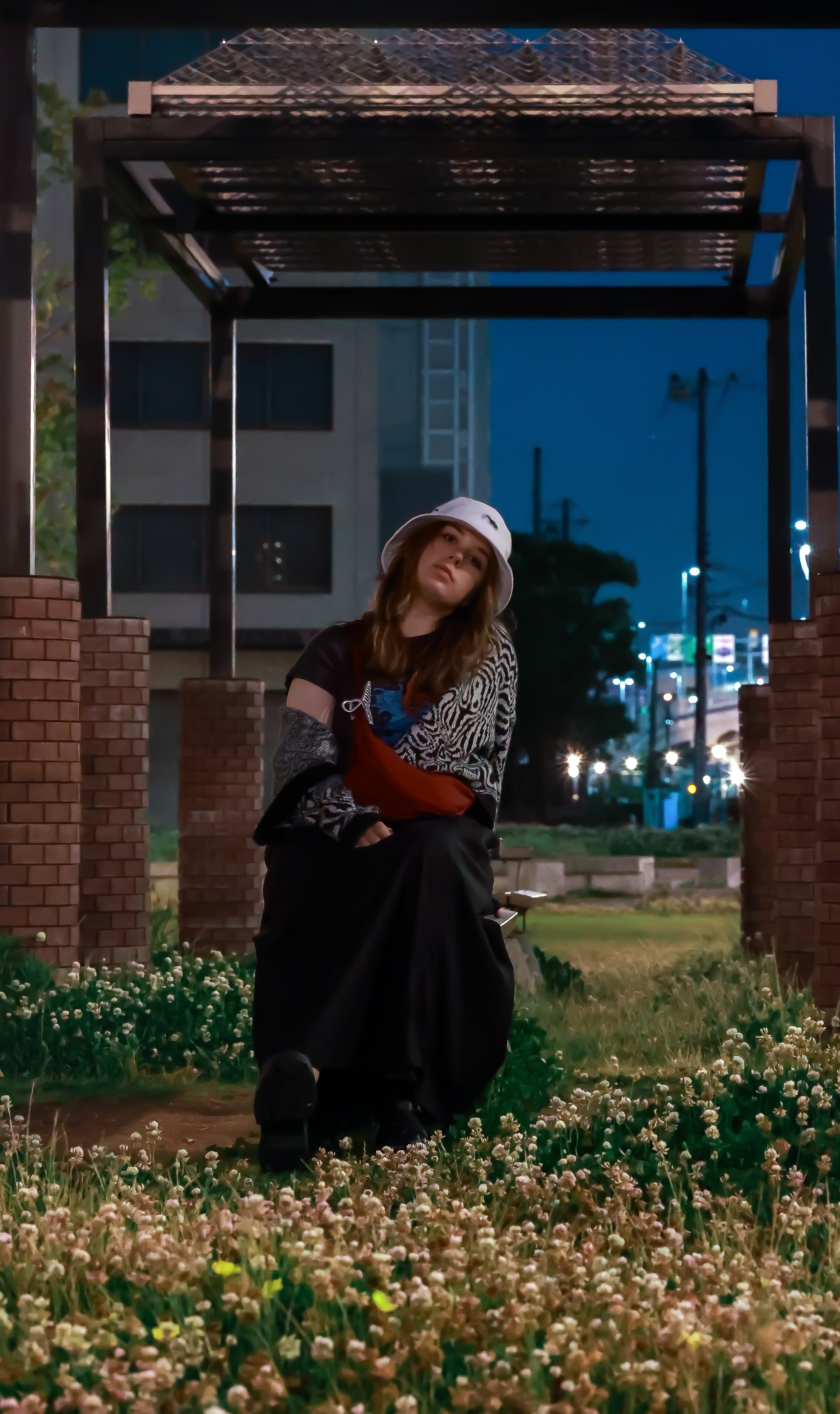 A woman wearing a white bucket hat, a black and patterned top, and black pants poses in a park at night. She is crouching on the grass surrounded by small white flowers, with an urban background that includes a building, streetlights, and parked cars