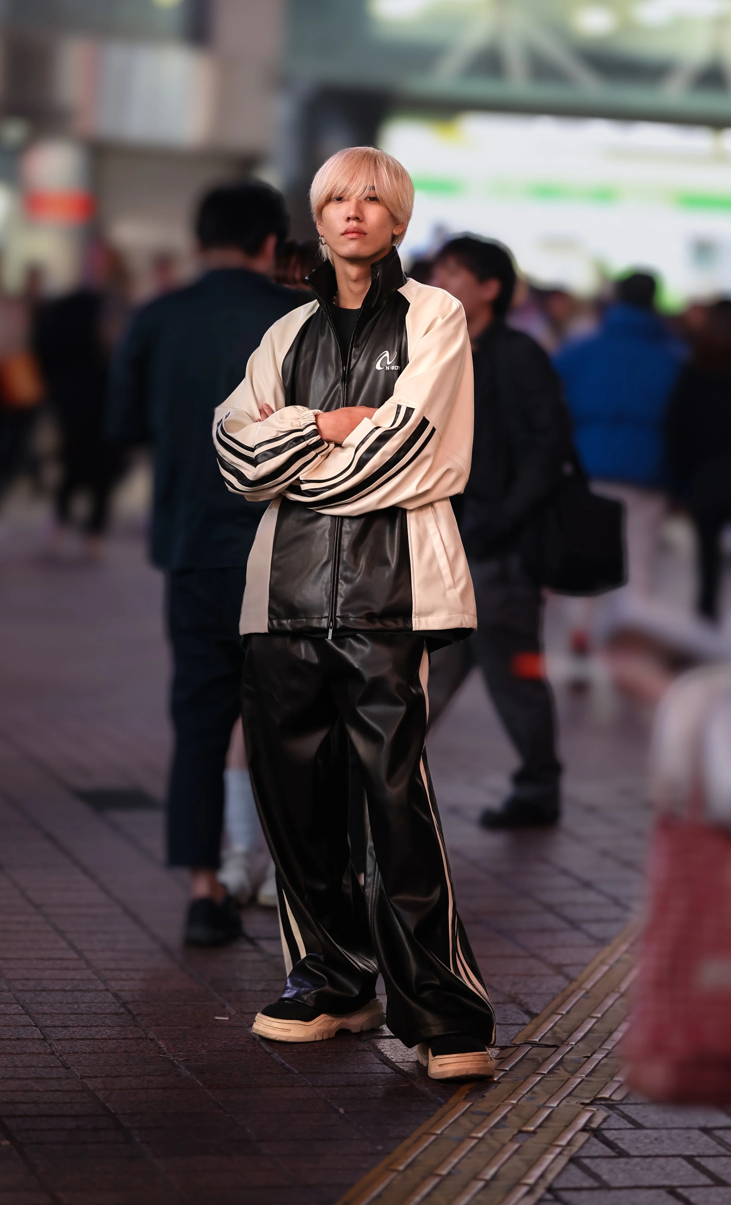 Young person with blonde hair standing with arms crossed in a busy indoor or outdoor space, wearing a black and white athletic jacket and black shiny pants, surrounded by other people.