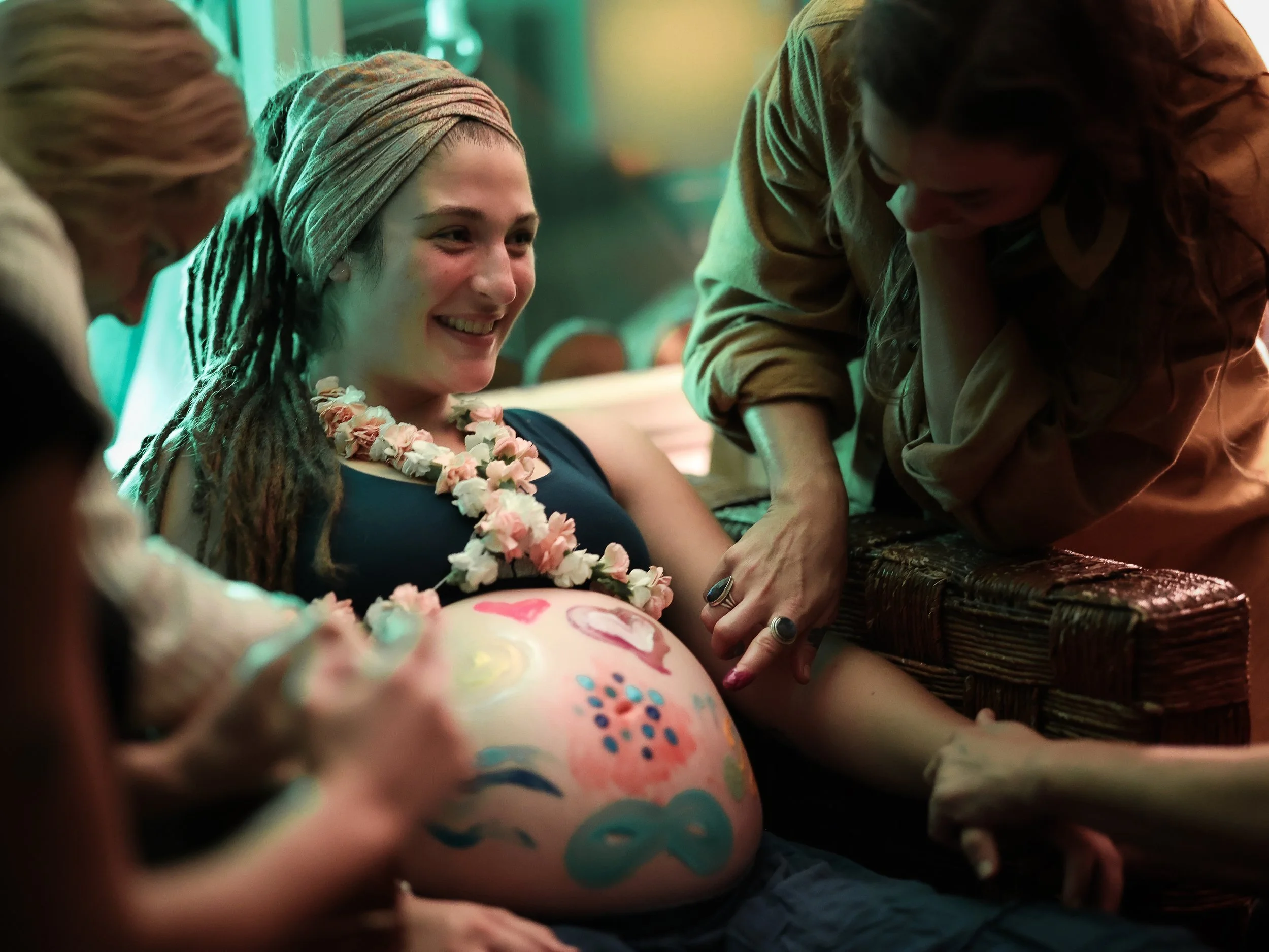 A woman with a headscarf and a flower necklace smiling and leaning over a pregnant woman's belly that is painted with colorful designs, surrounded by people at a celebration or gathering.