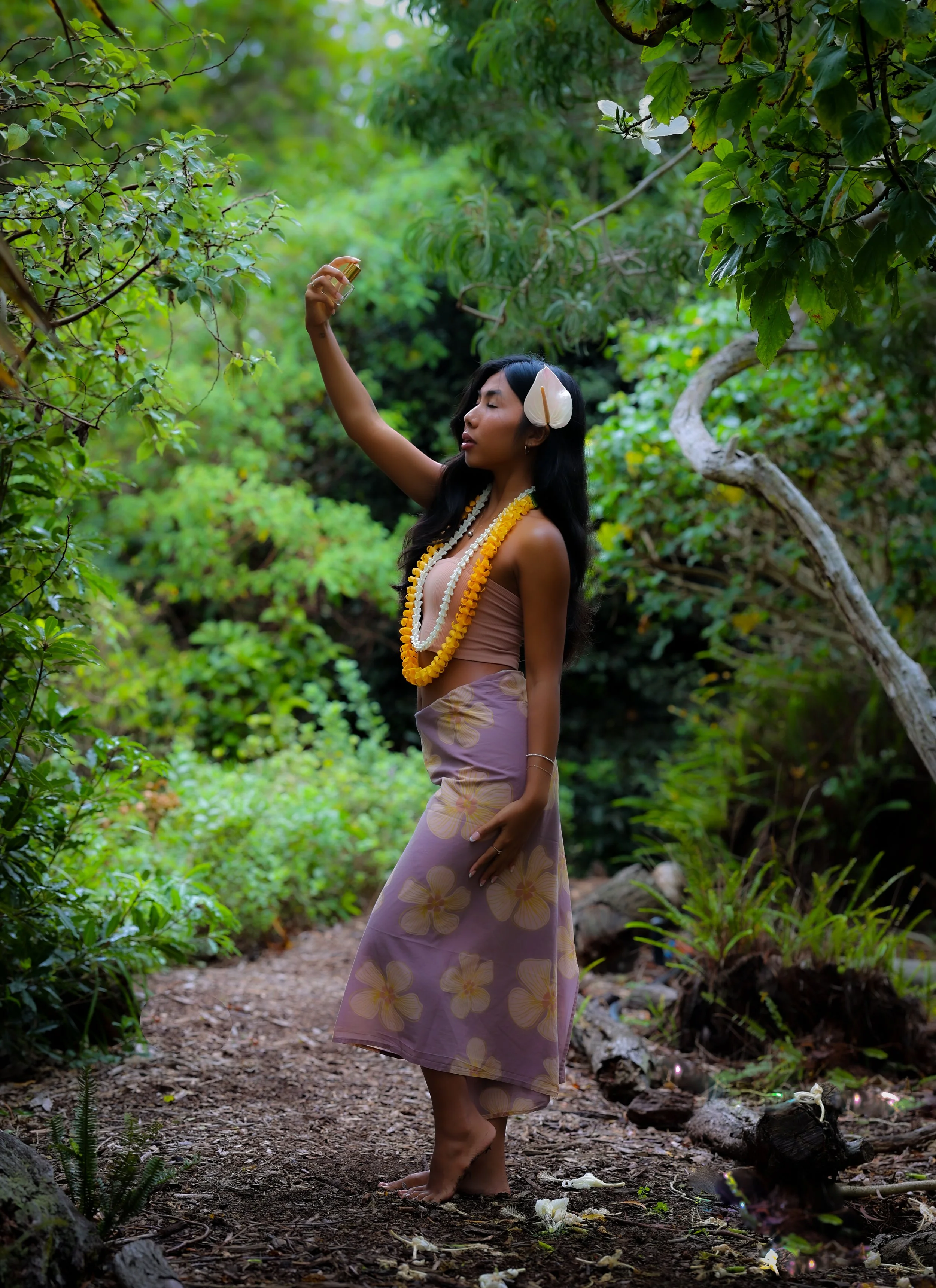 A woman standing barefoot on a forest trail, wearing a light brown top and a floral skirt, with white flowers and yellow leis around her neck, raising her hand holding a small bottle in a lush, green, tropical setting.